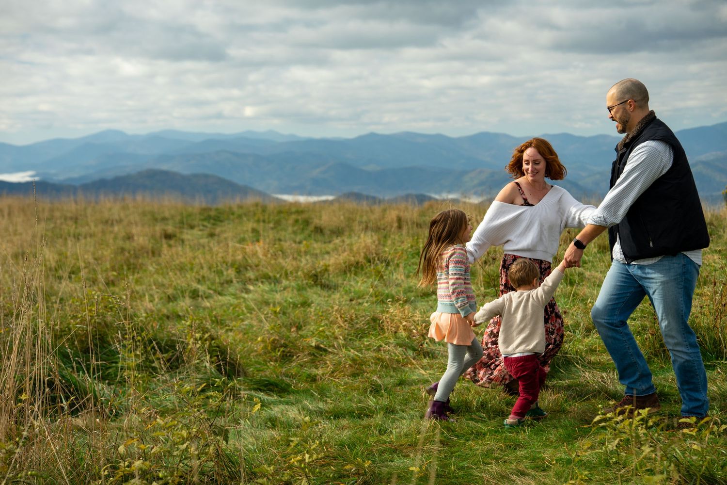 Family members sit together in a grassy meadow overlooking mountain views on a cloudy summer day.