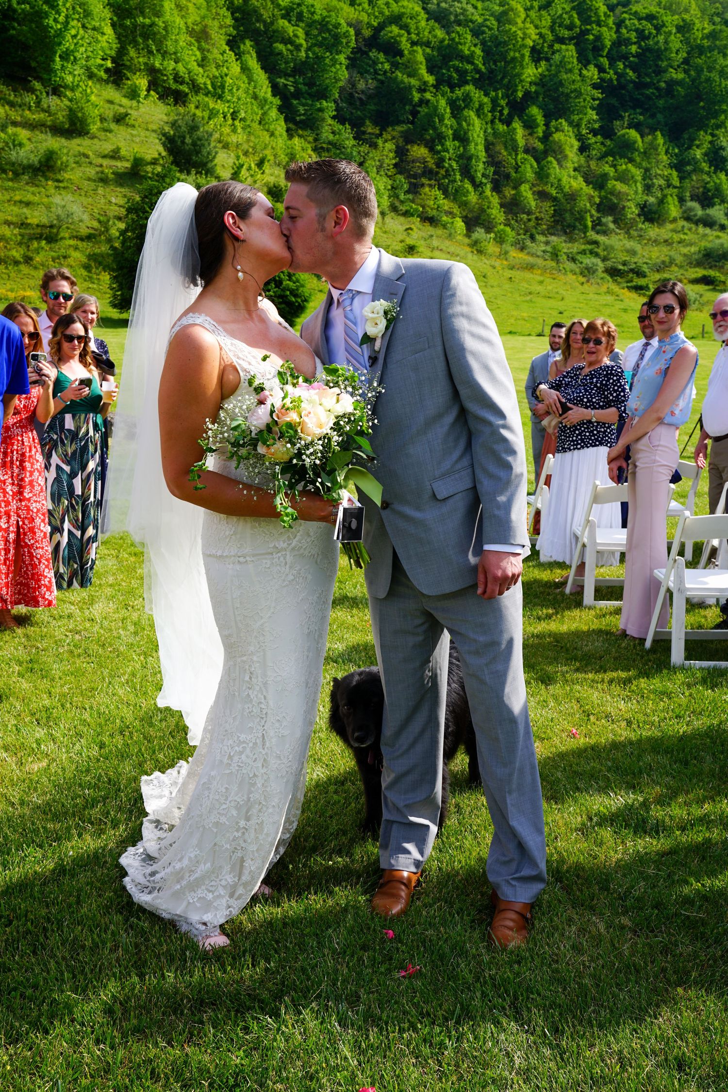 A romantic outdoor wedding ceremony shows a couple sharing their first kiss as newlyweds on a lush green hillside.