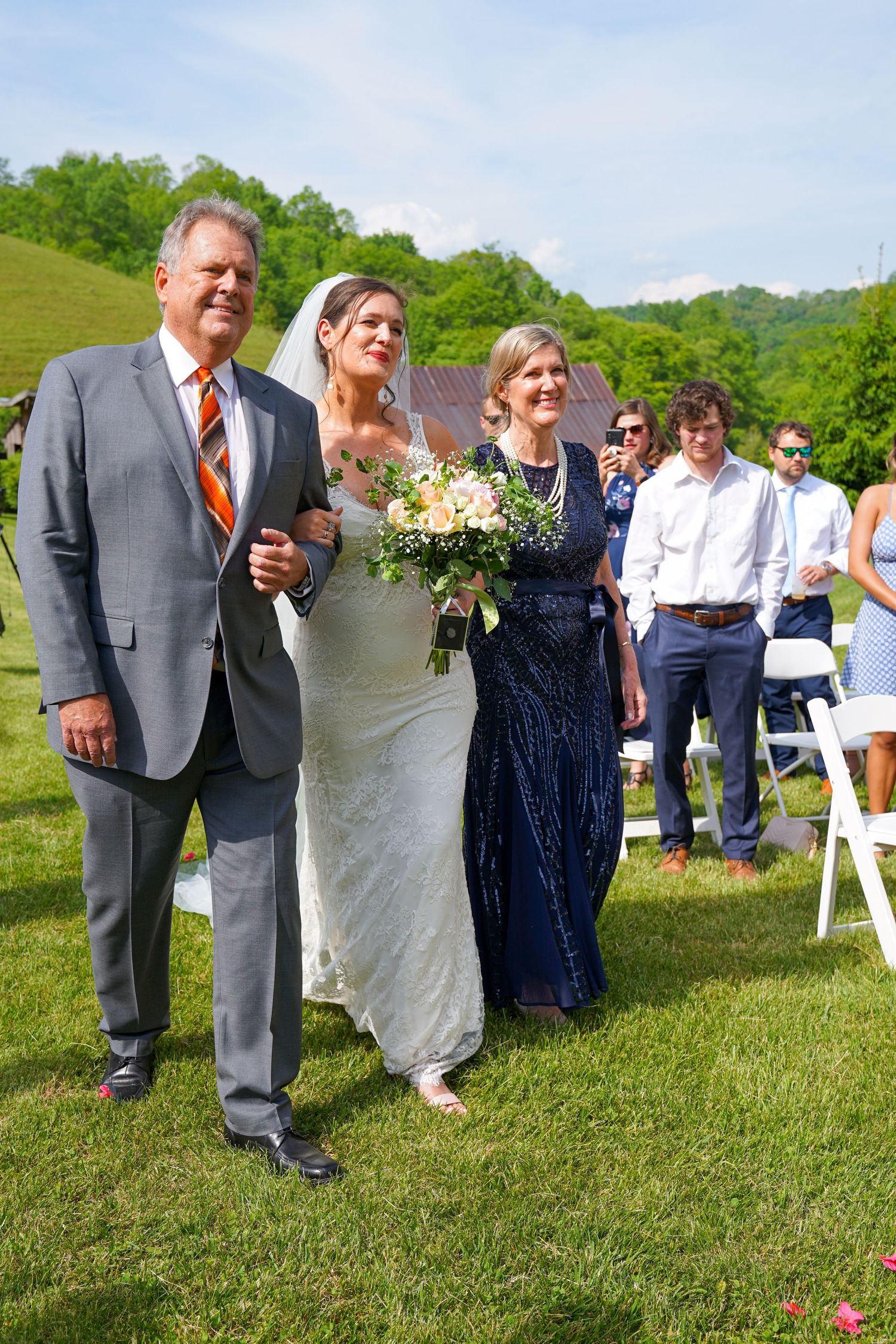 A group of wedding guests walk in a line across a grassy lawn at an outdoor ceremony on a sunny day.