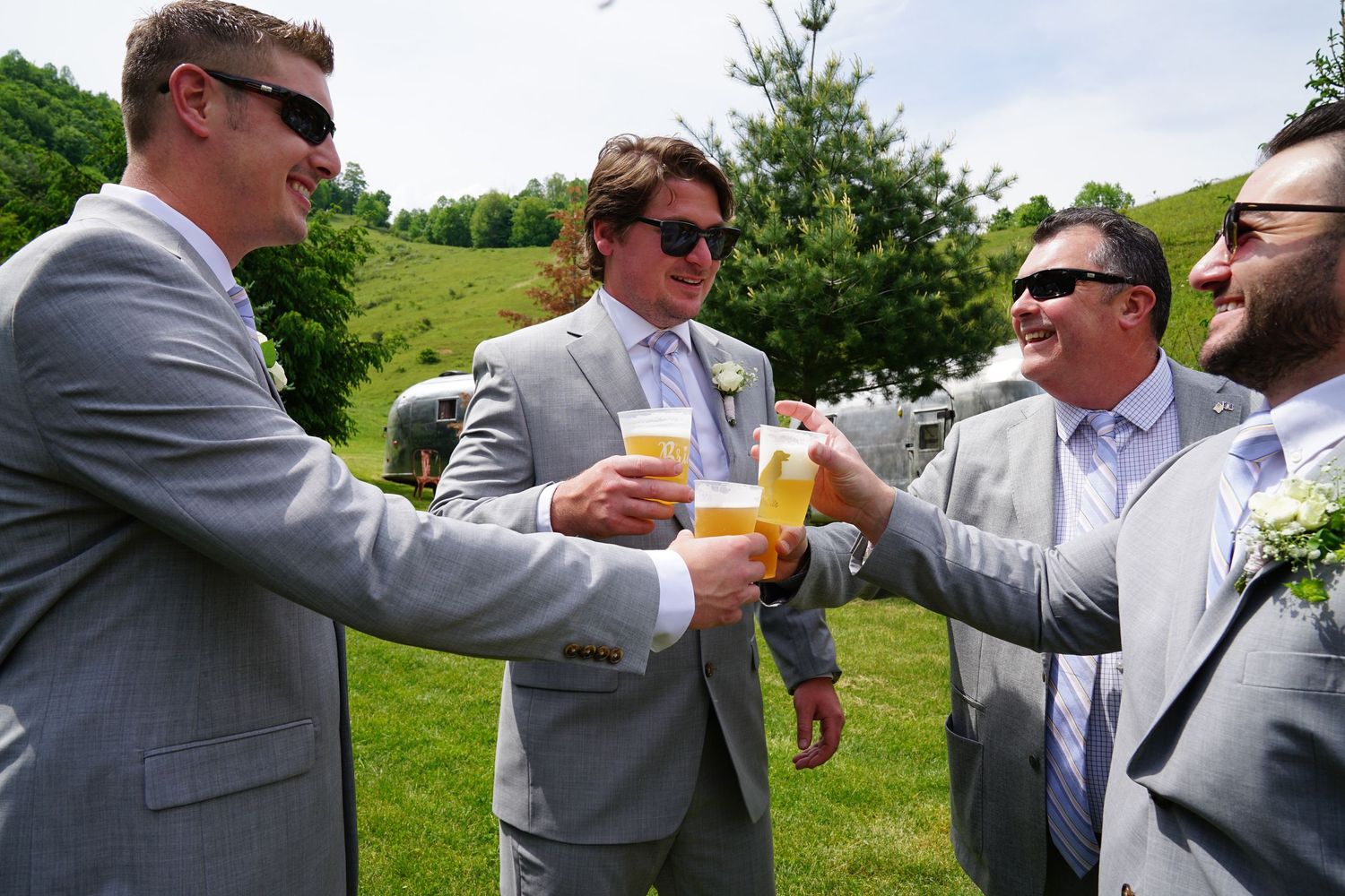 A group of groomsmen in gray suits share drinks and toast together during an outdoor celebration.