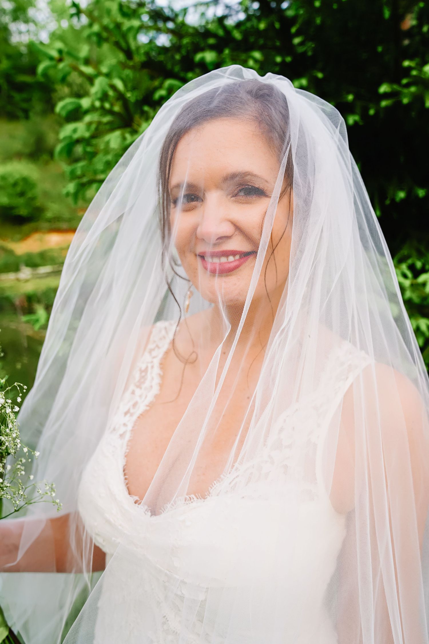 A bride wearing a white wedding dress and long veil smiles while standing in front of green foliage outdoors.