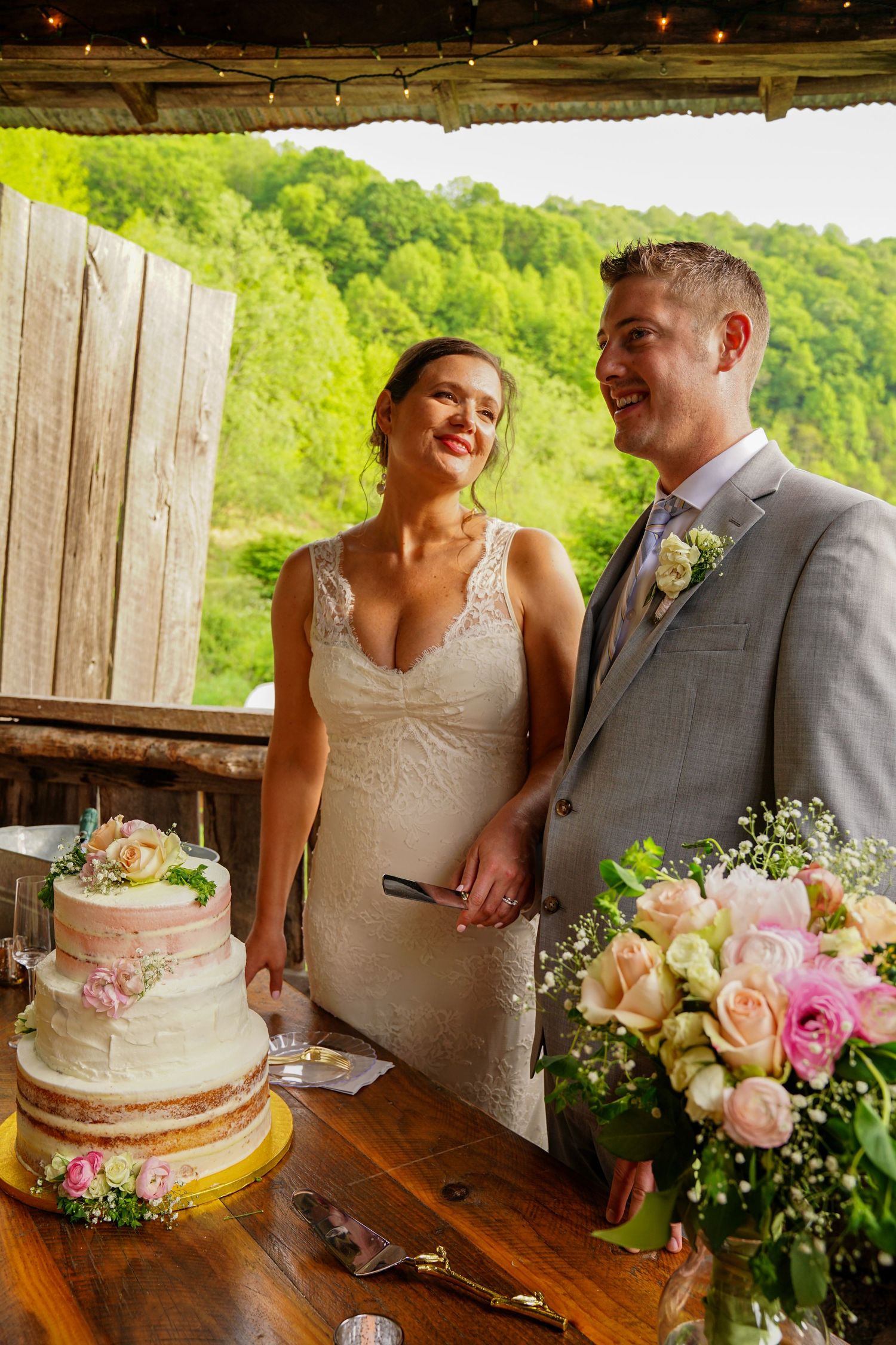 A happy couple shares a laugh while standing next to their pink and white wedding cake and floral bouquet at a rustic venue.