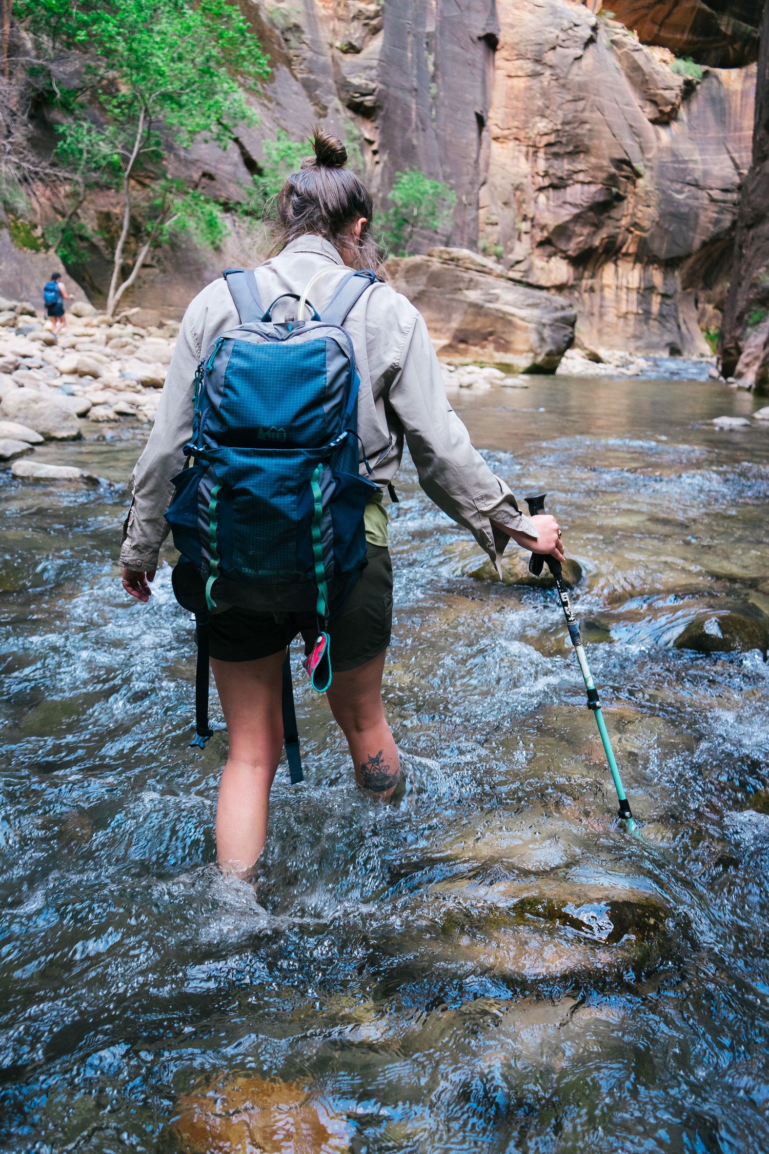 A hiker wearing blue backpack wading through a rocky stream in a narrow canyon while holding hiking poles.