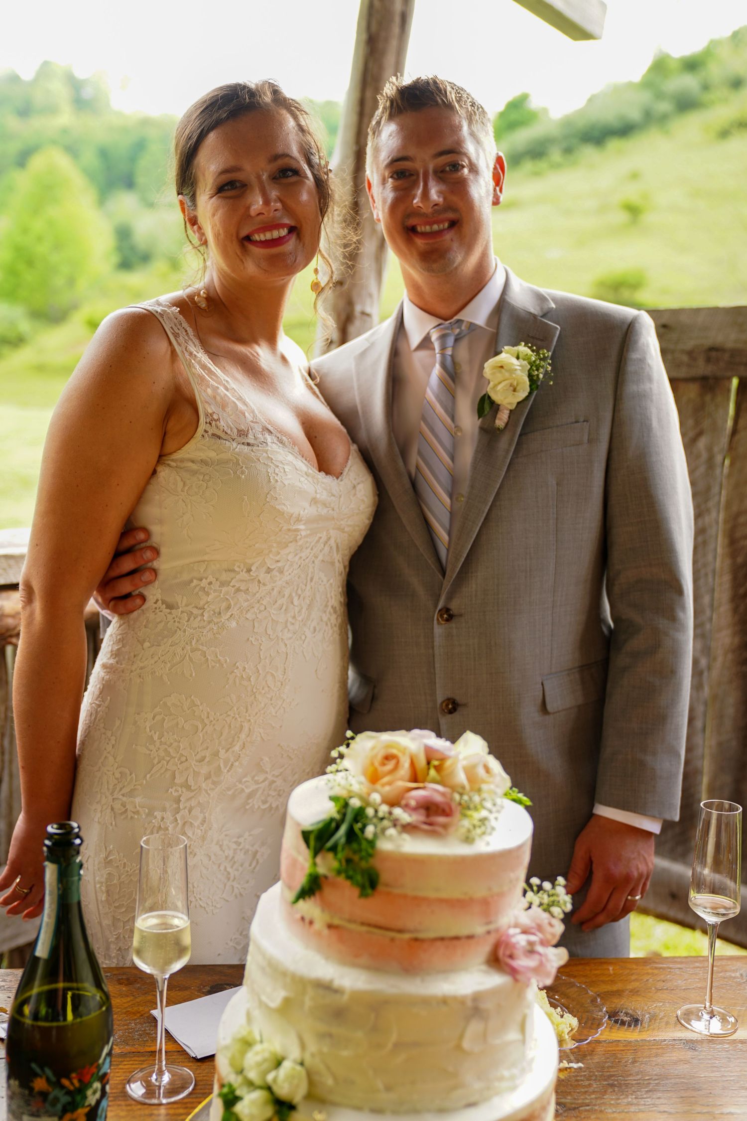 A wedding couple cuts into their simple white buttercream cake decorated with pink roses during an outdoor celebration.