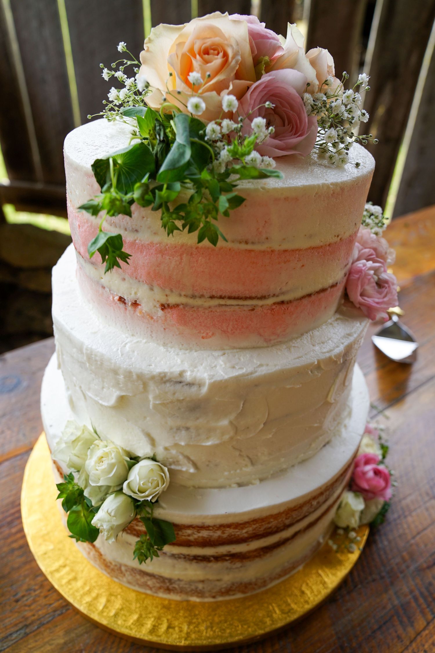 A rustic semi-naked wedding cake decorated with fresh pink and peach roses and greenery on a gold cake board.