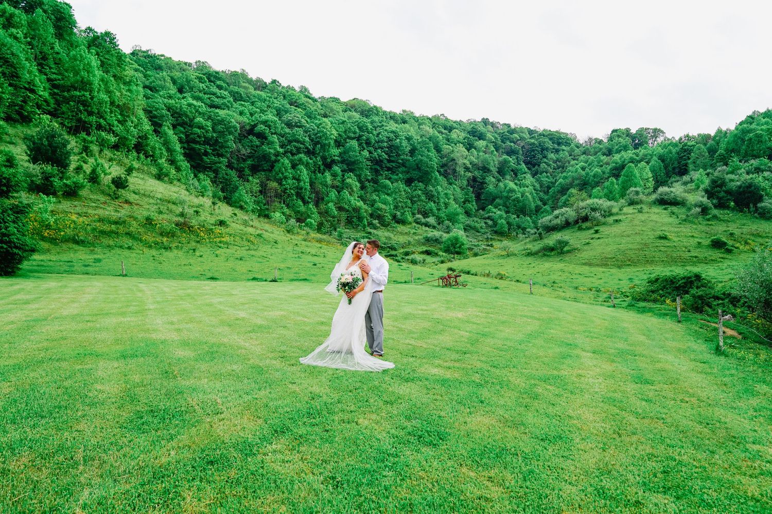 A couple in wedding attire stands together on a lush green hillside surrounded by vibrant forest scenery.