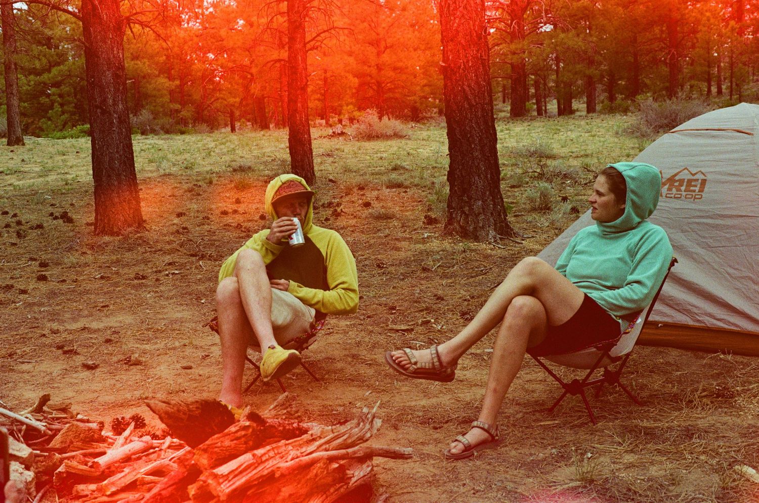 Two campers relax at a campsite during sunset with tents and camping gear in a wooded area.