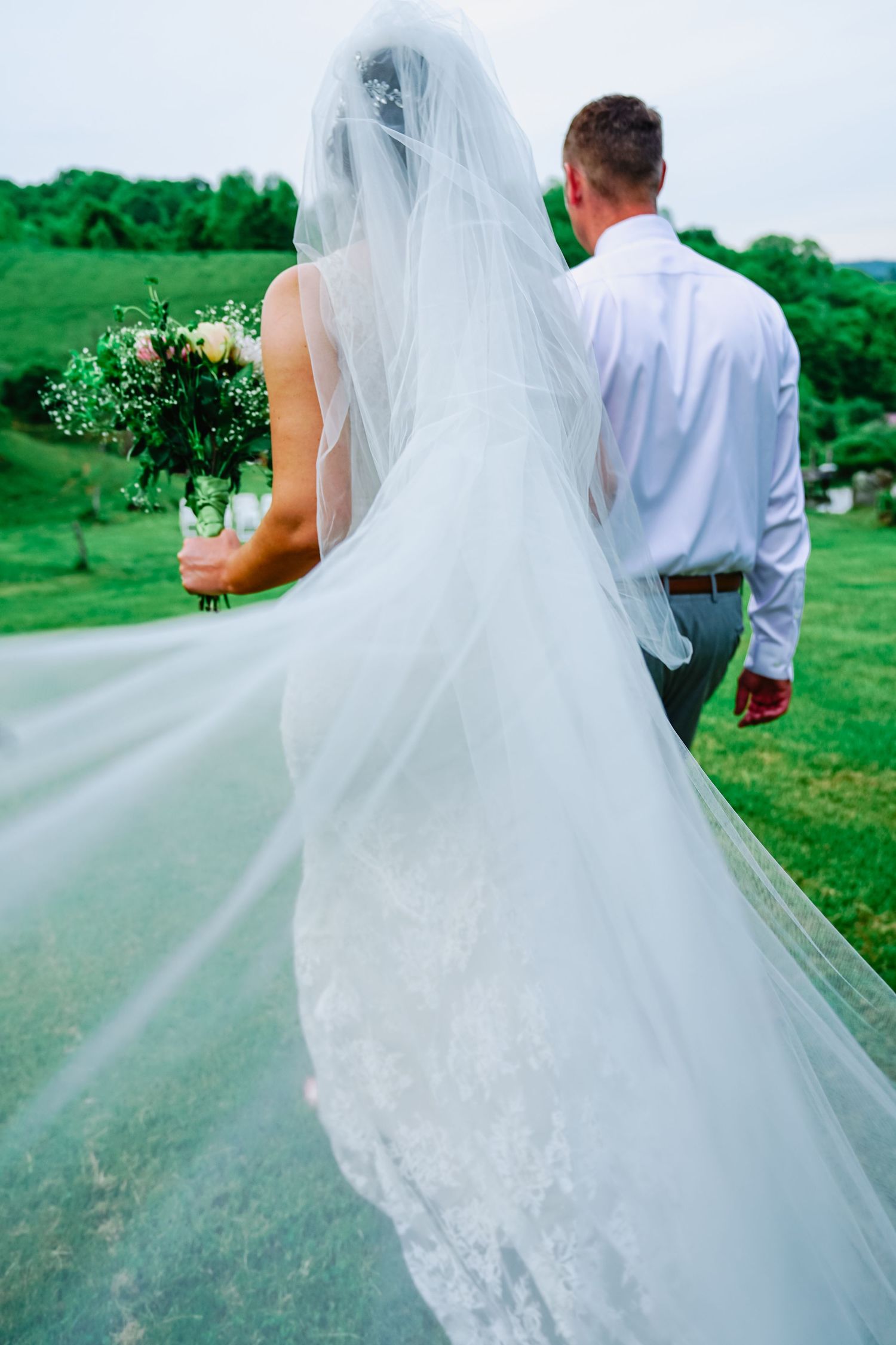 A flowing white wedding veil trails behind a couple walking on a green hillside in a romantic outdoor setting.