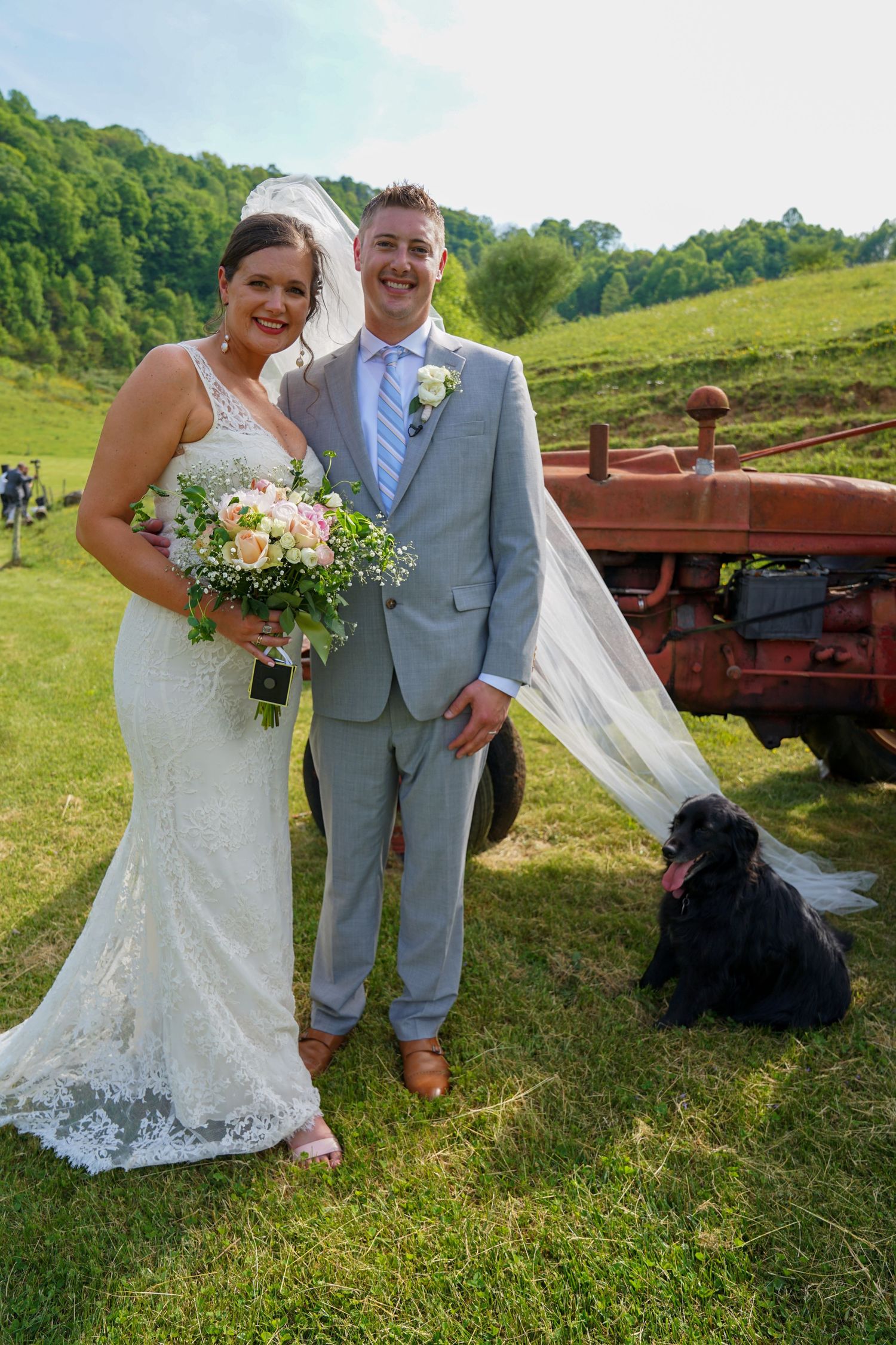 A couple poses for wedding photos in a rustic countryside setting with a vintage red farm wagon in the background.