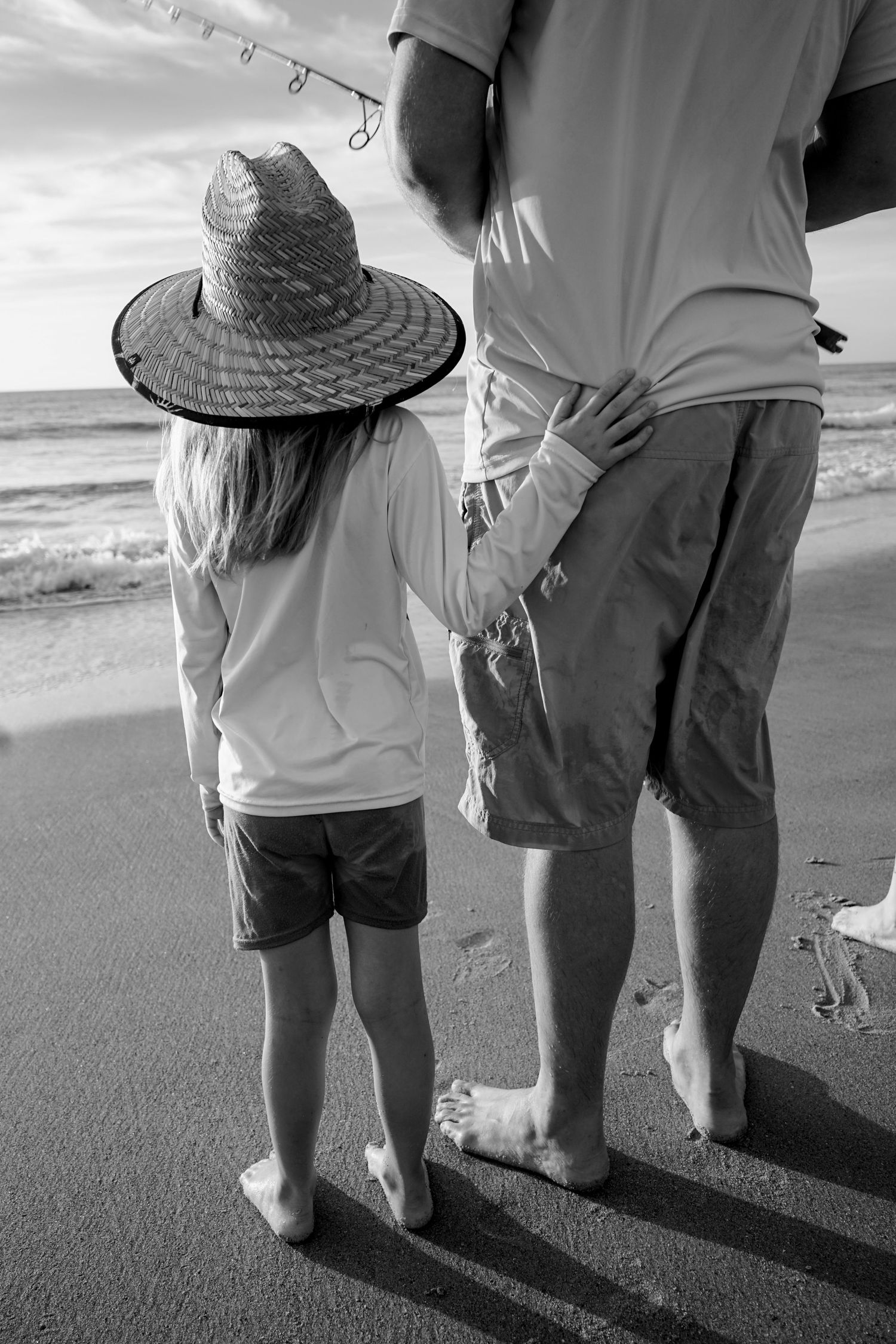 Black and white image of figures standing on beach with fishing poles and straw hat.