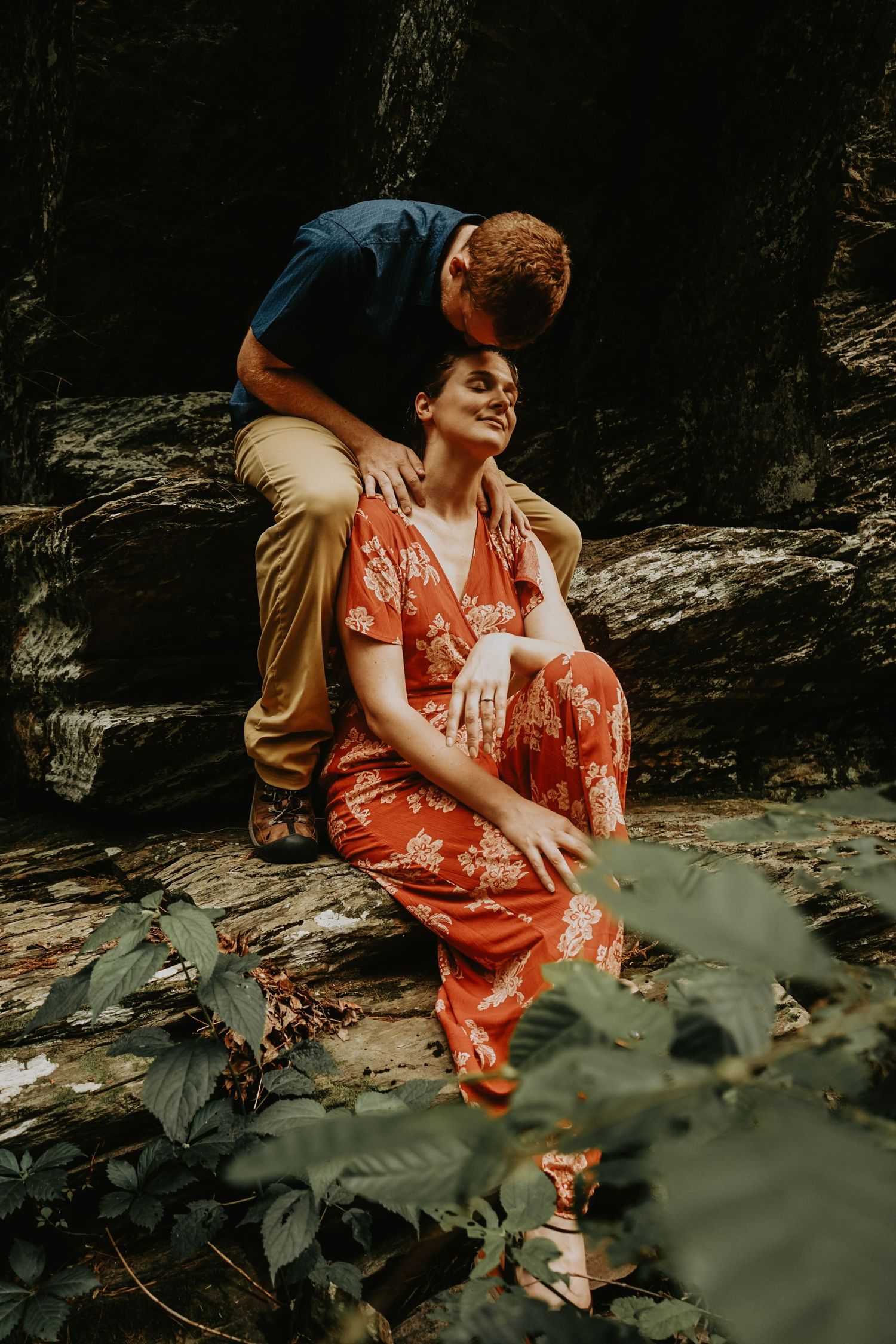 A couple posing intimately on rocky terrain surrounded by lush greenery in moody outdoor lighting.