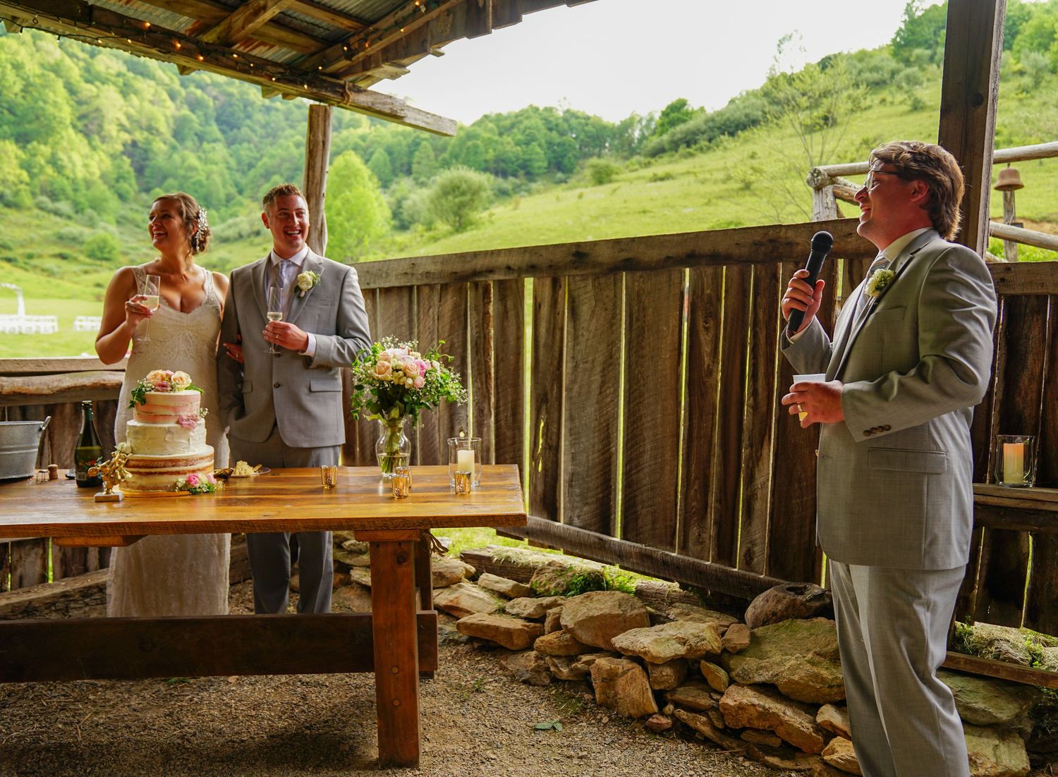 People gather in a rustic wooden barn structure with tables and benches set up for an event or gathering.
