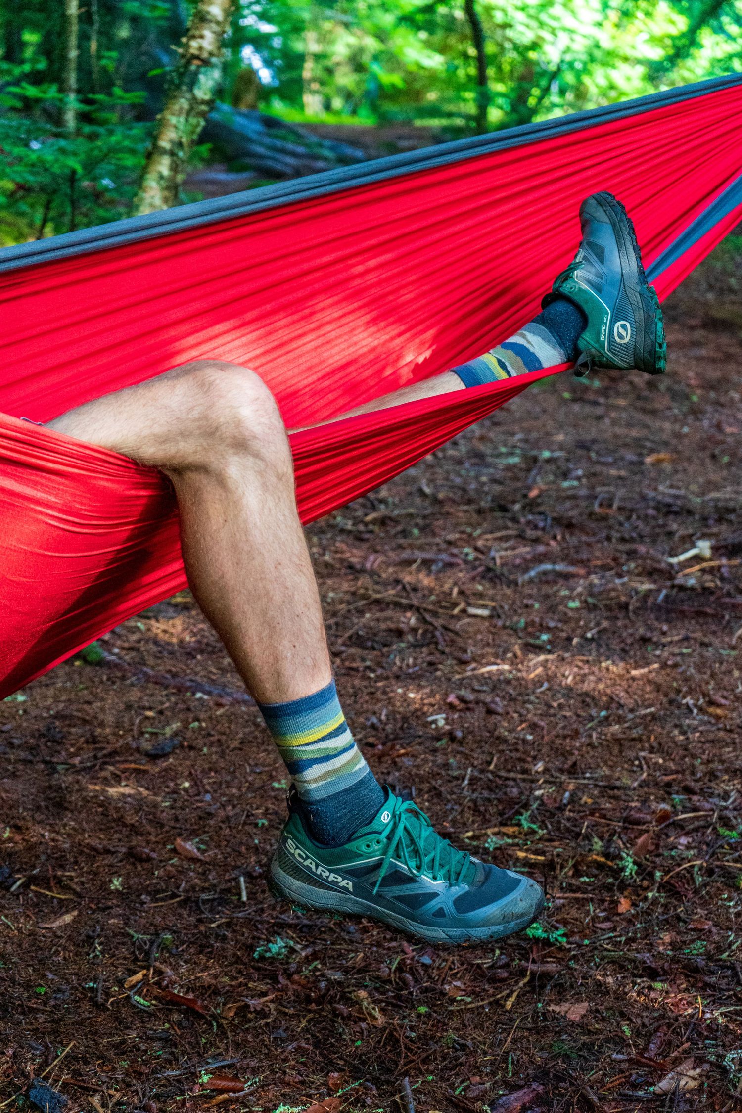 Close-up view of green hiking shoes in a red hammock against forest ground.