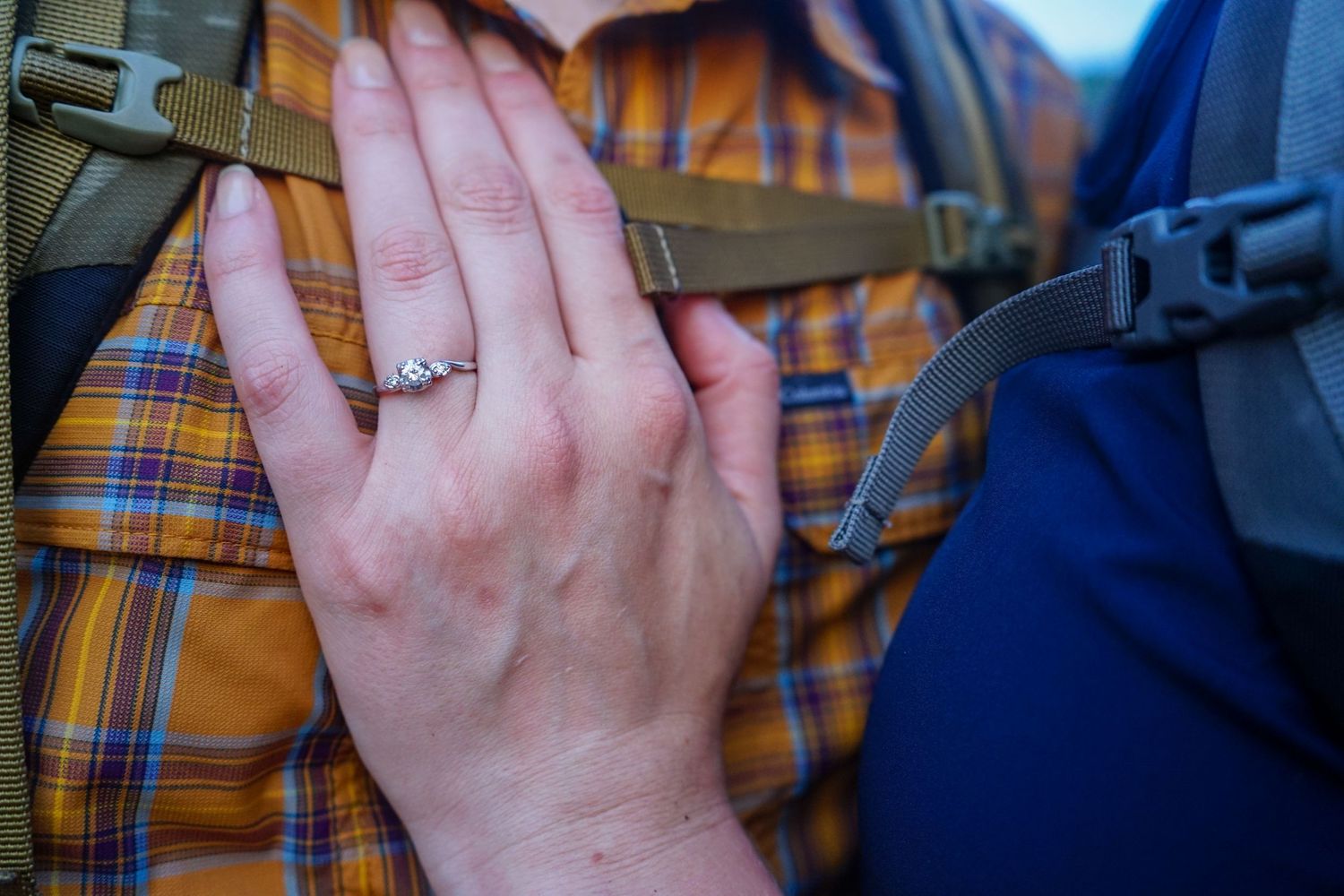 Hand wearing a diamond ring shown against a plaid orange shirt and blue backpack straps in close-up.