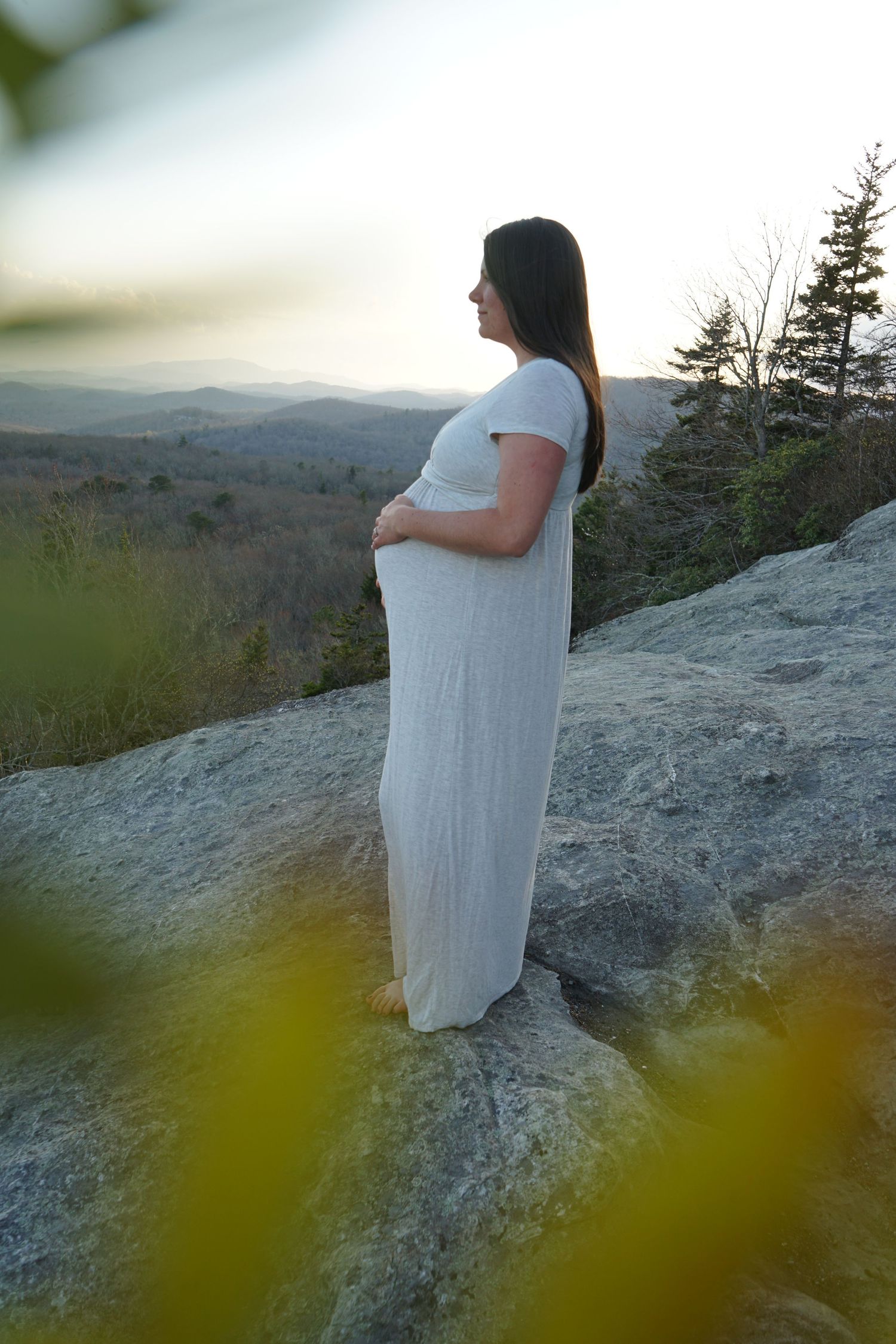 A pregnant woman in a white dress stands on a rocky overlook admiring a mountain vista.