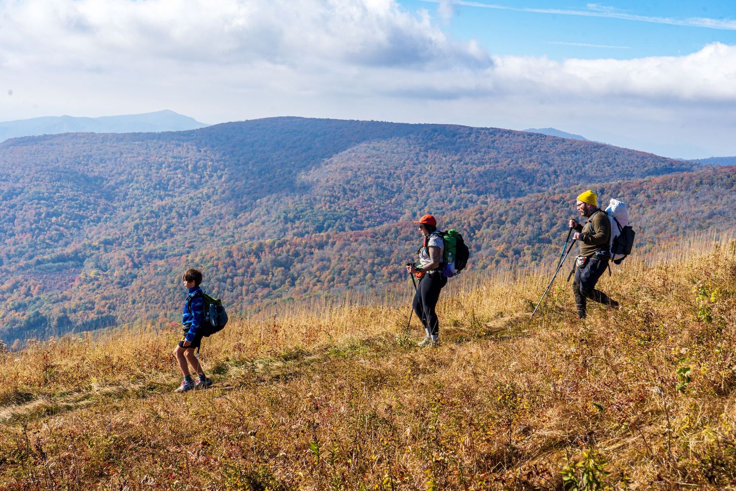Hikers traverse a scenic mountain ridge trail with rolling autumn hills in the background.