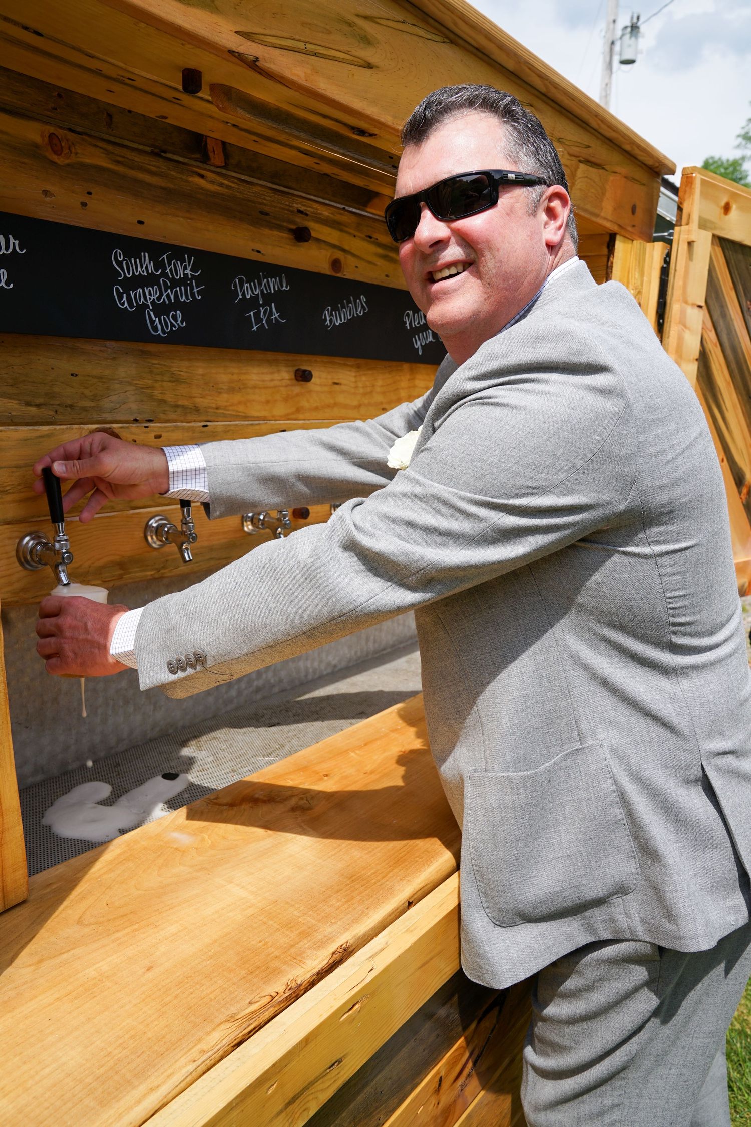 A person in a gray suit writes on a chalkboard menu mounted on a wooden wall at an outdoor venue.
