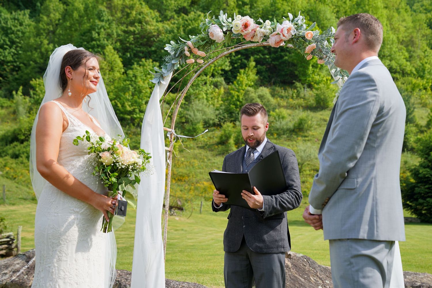 A wedding ceremony takes place outdoors with white floral arrangements and a draped arch in a lush green garden setting.
