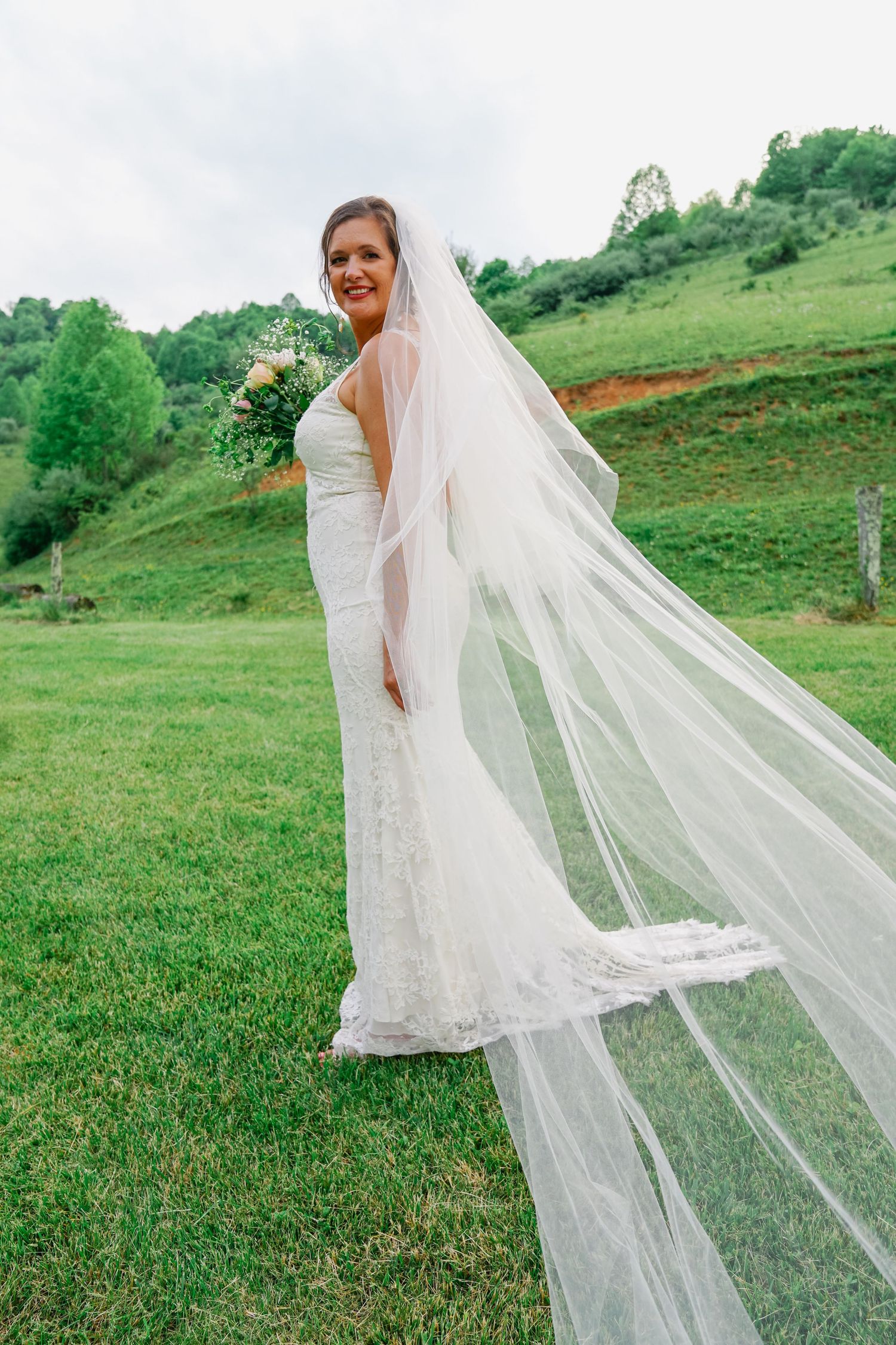A bride in a white lace wedding dress stands in a green field with her flowing veil caught in the breeze.