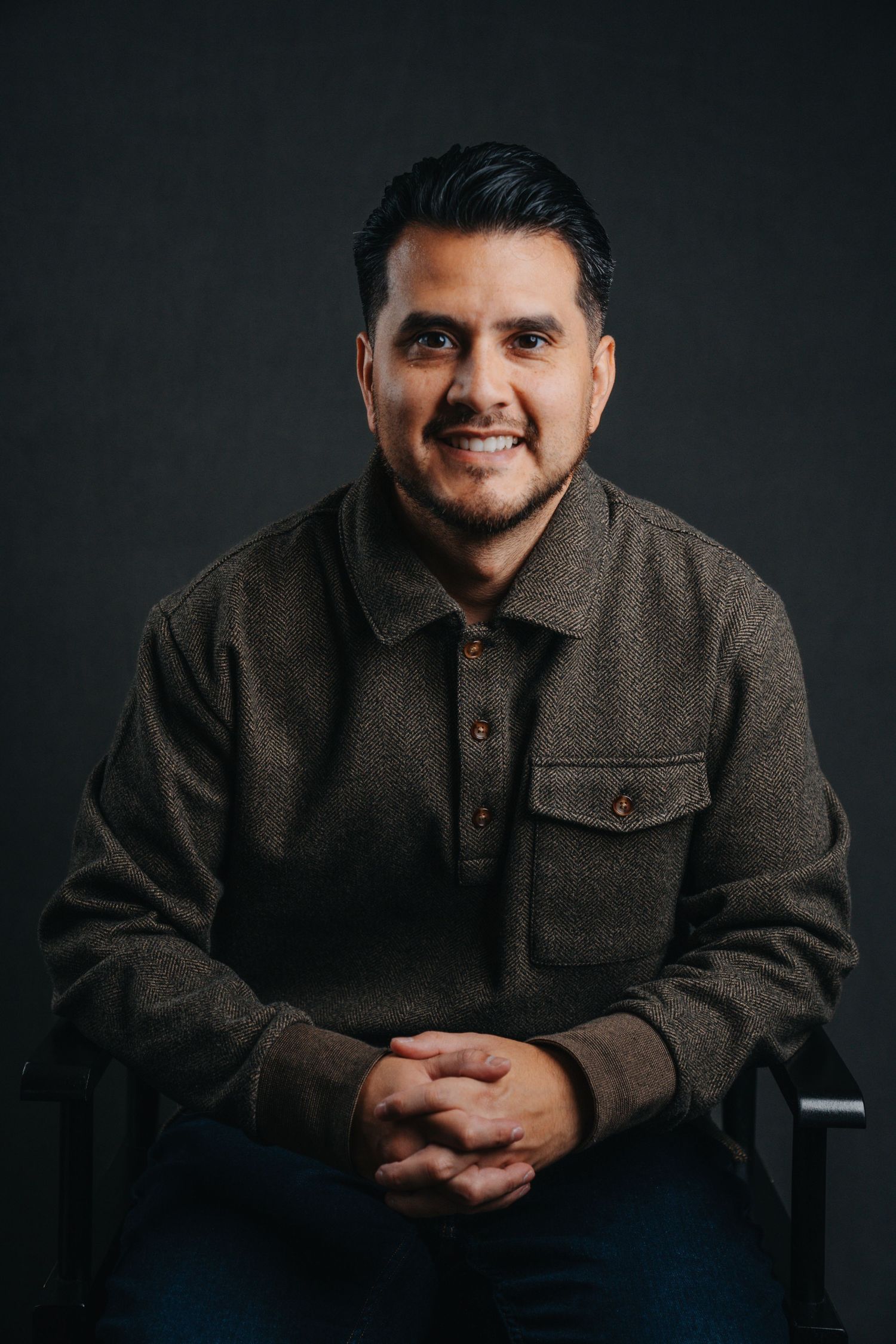 Smiling man with dark hair and beard, wearing a brown button-up shirt, seated with hands clasped against dark background.