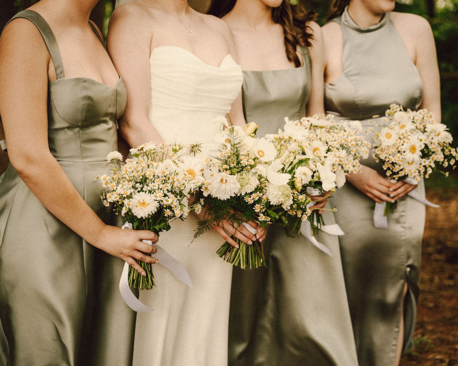 Bride and bridesmaids in sage green satin dresses holding white floral bouquets at outdoor wedding.
