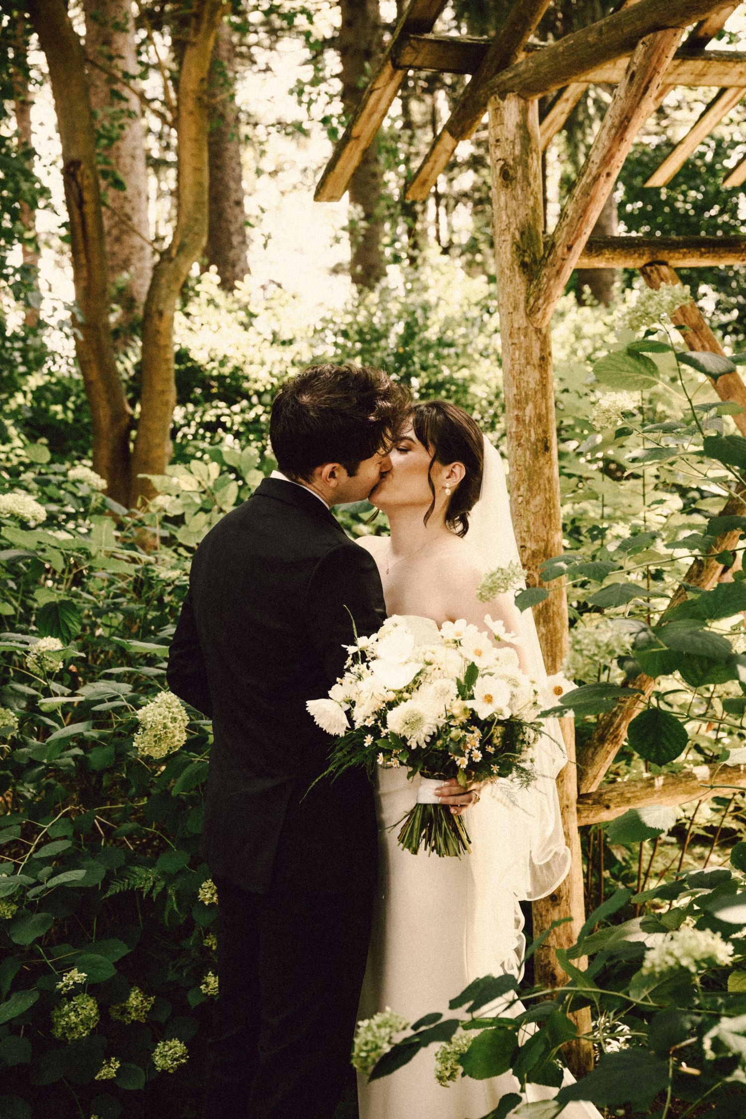 Bride and groom share a tender kiss beneath a rustic wooden pergola surrounded by lush green hydrangeas.