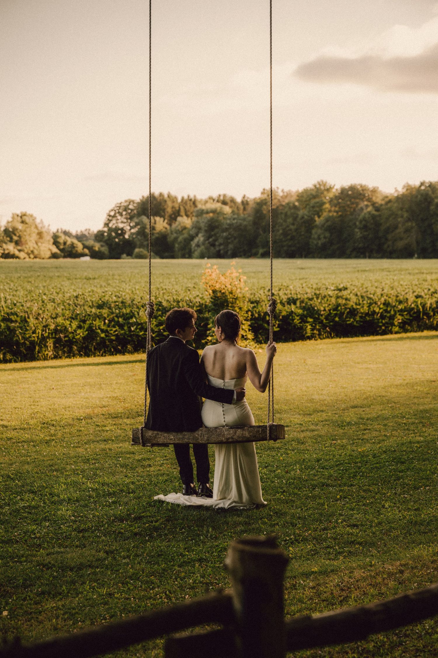Bride and groom sitting together on wooden swing overlooking vineyard at sunset.