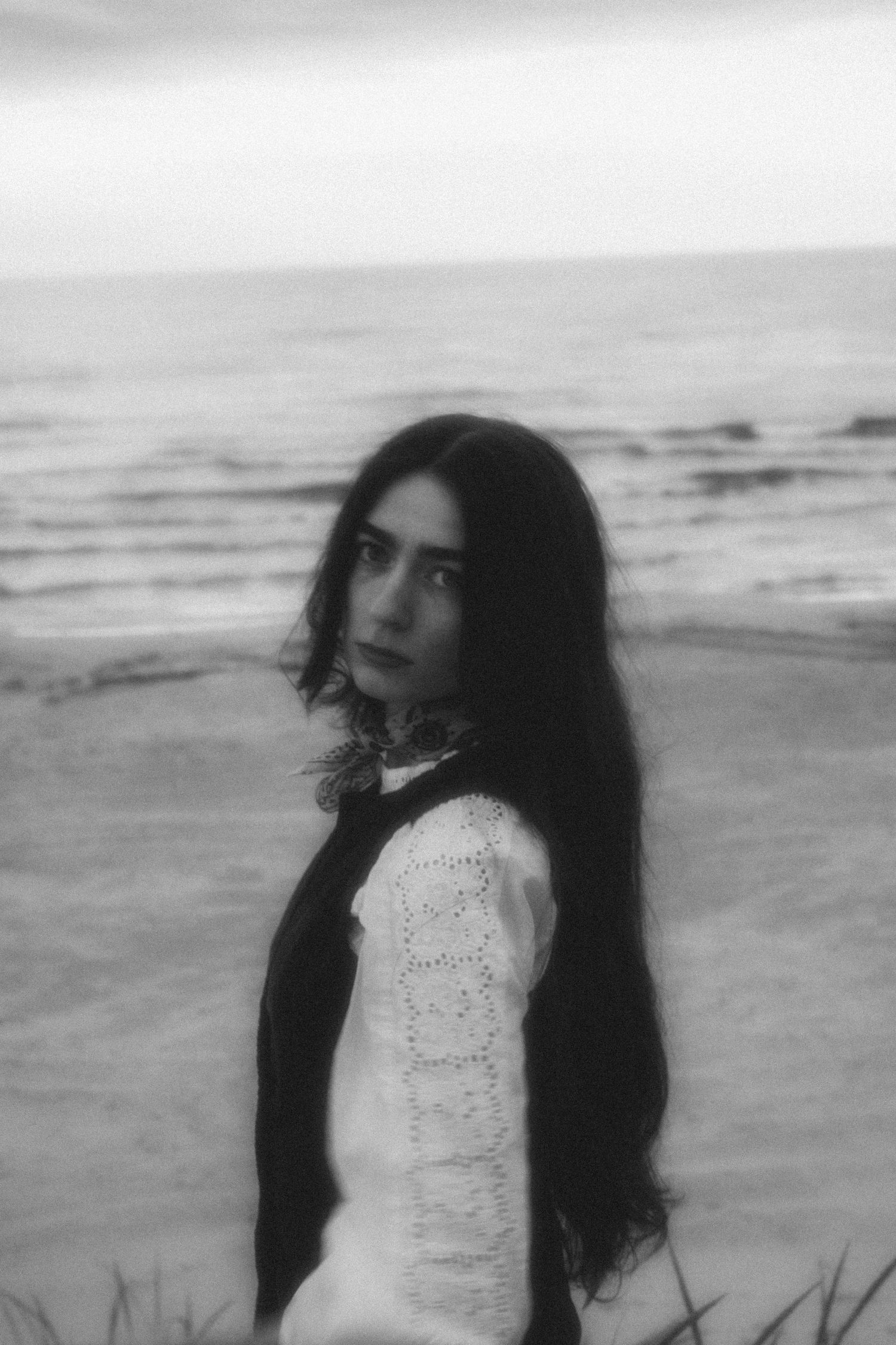 Black and white portrait of dark-haired woman in white lace top standing on beach.