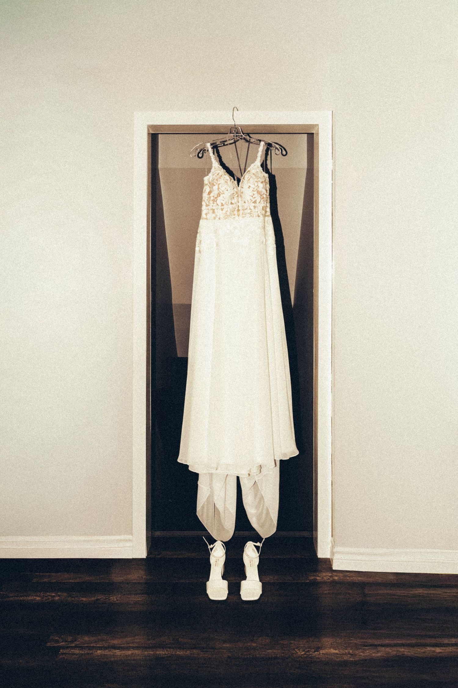 White lace wedding dress with white strappy heels hanging in a doorframe on dark hardwood floor.