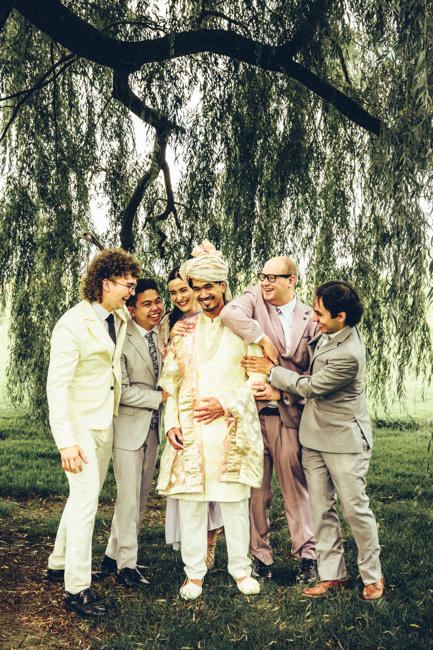 Groom in traditional Indian sherwani surrounded by groomsmen in suits under weeping willow tree.