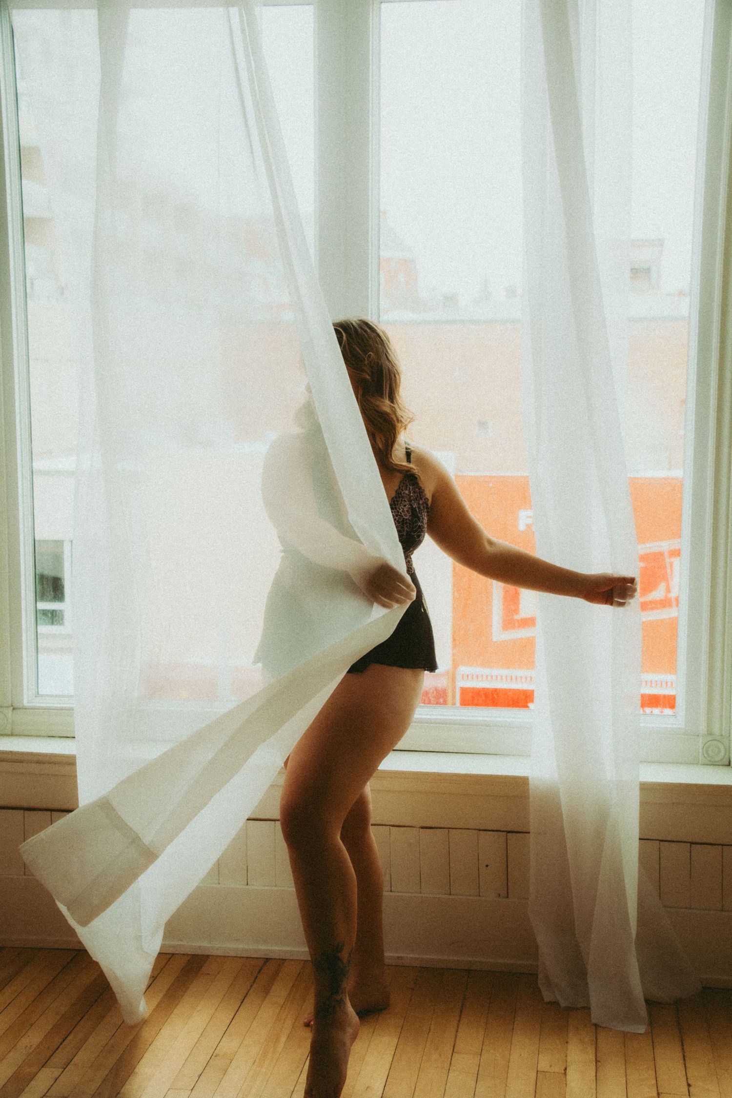 Woman in black lingerie standing by sheer white curtains with city view in warm natural light.