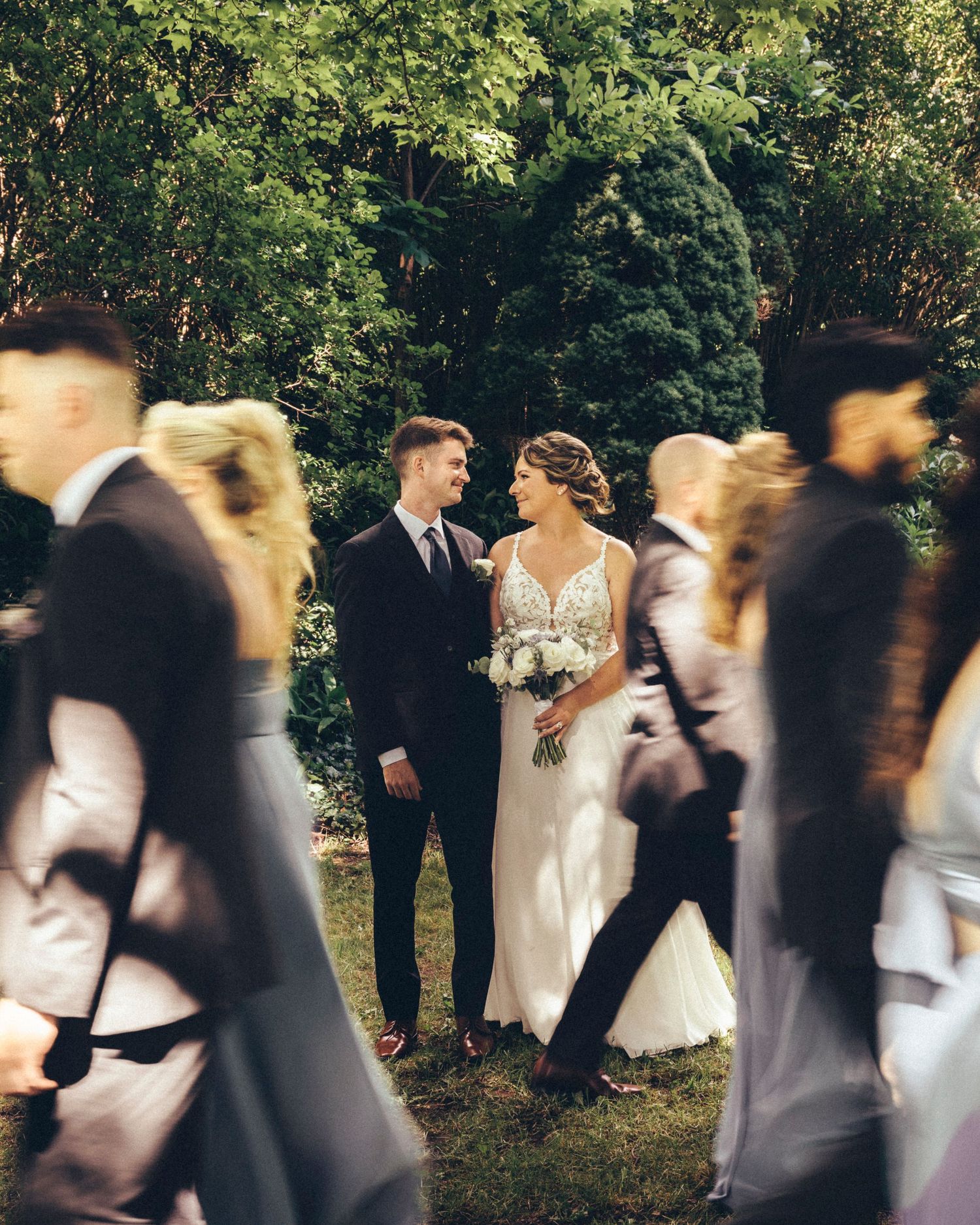 Bride and groom gazing at each other as blurred wedding guests walk past in garden.