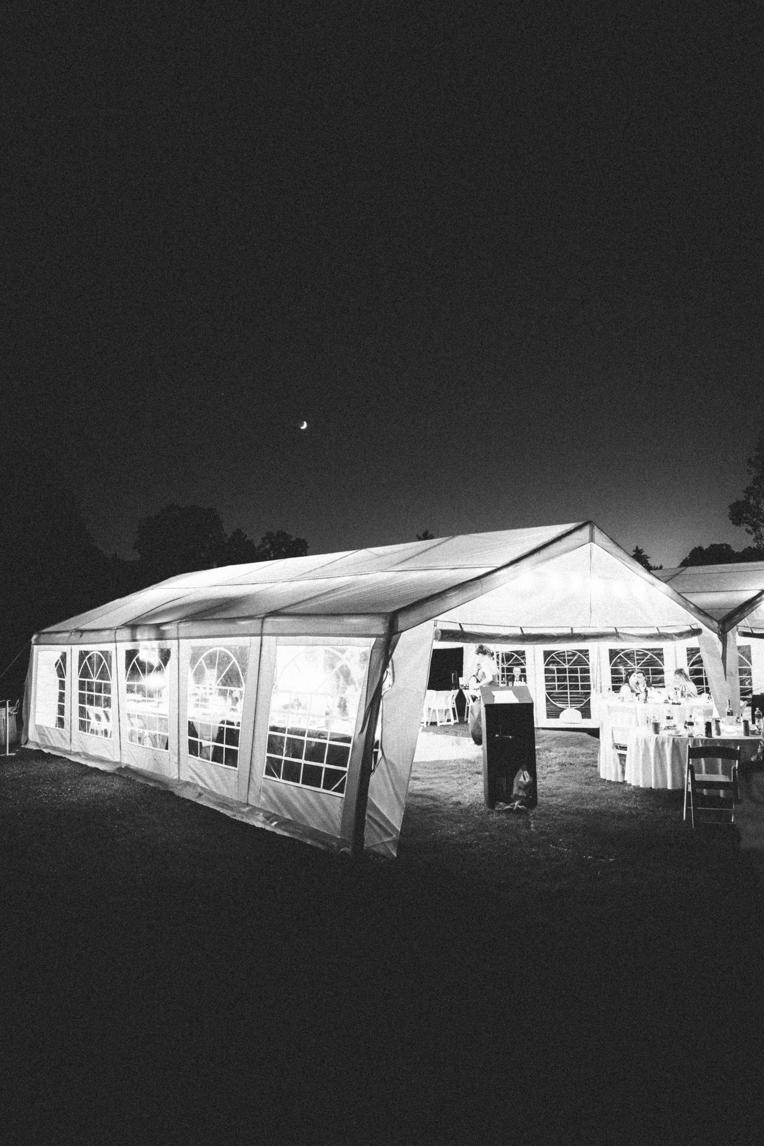 Illuminated white wedding reception tent glows against dark night sky with crescent moon.