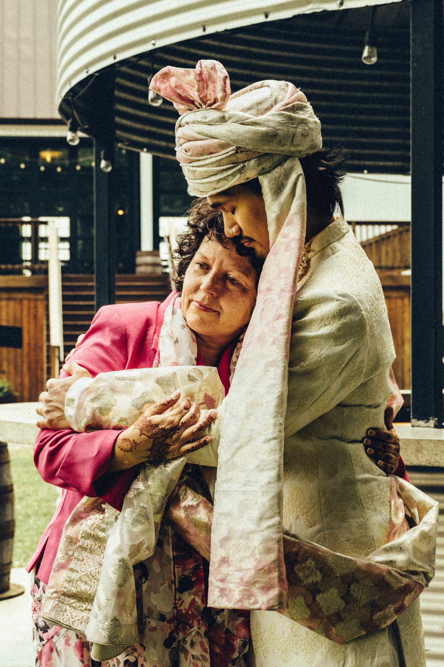 Emotional wedding moment as an older woman in pink embraces a groom in traditional South Asian attire with a floral turban.
