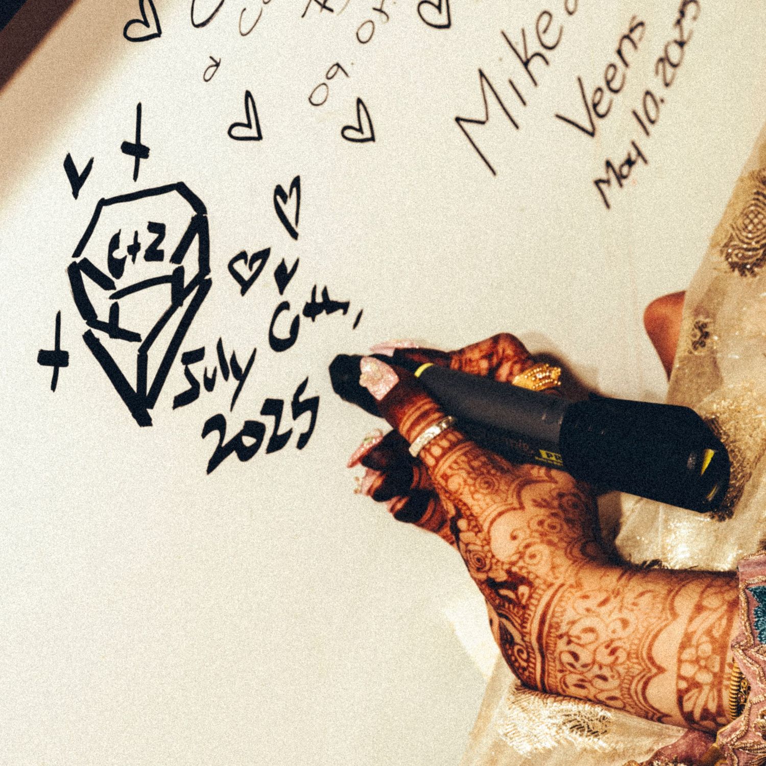 Henna-adorned hand writing wedding date July 6th 2025 on a white guest signing board.