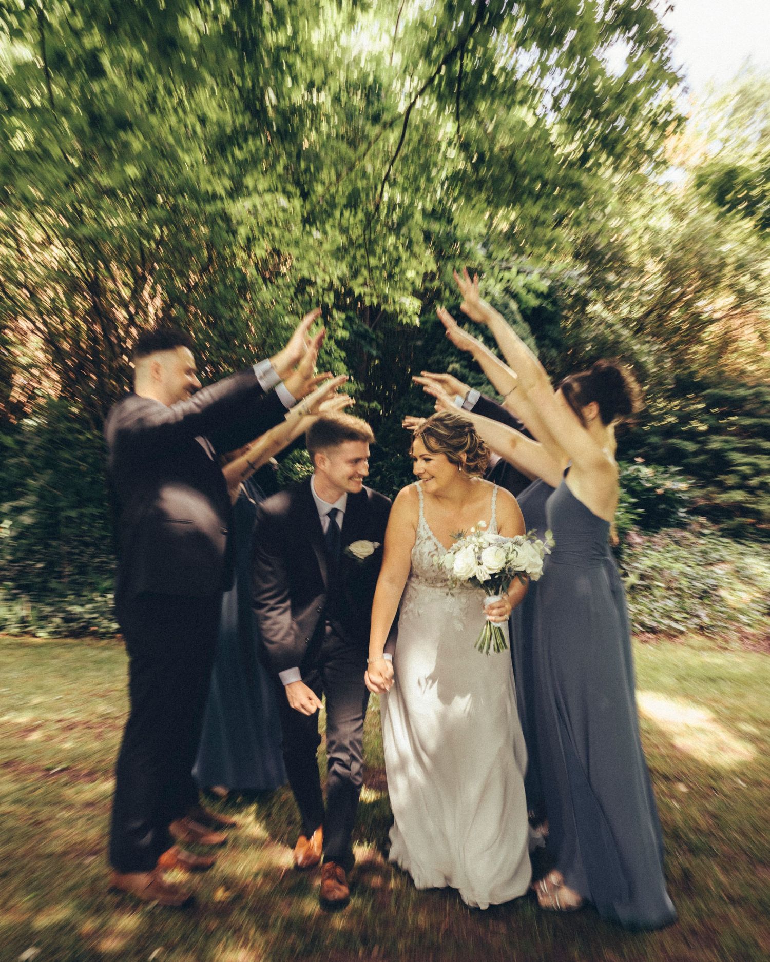 Bride and groom walk hand-in-hand as wedding party raises arms in celebration outdoors.