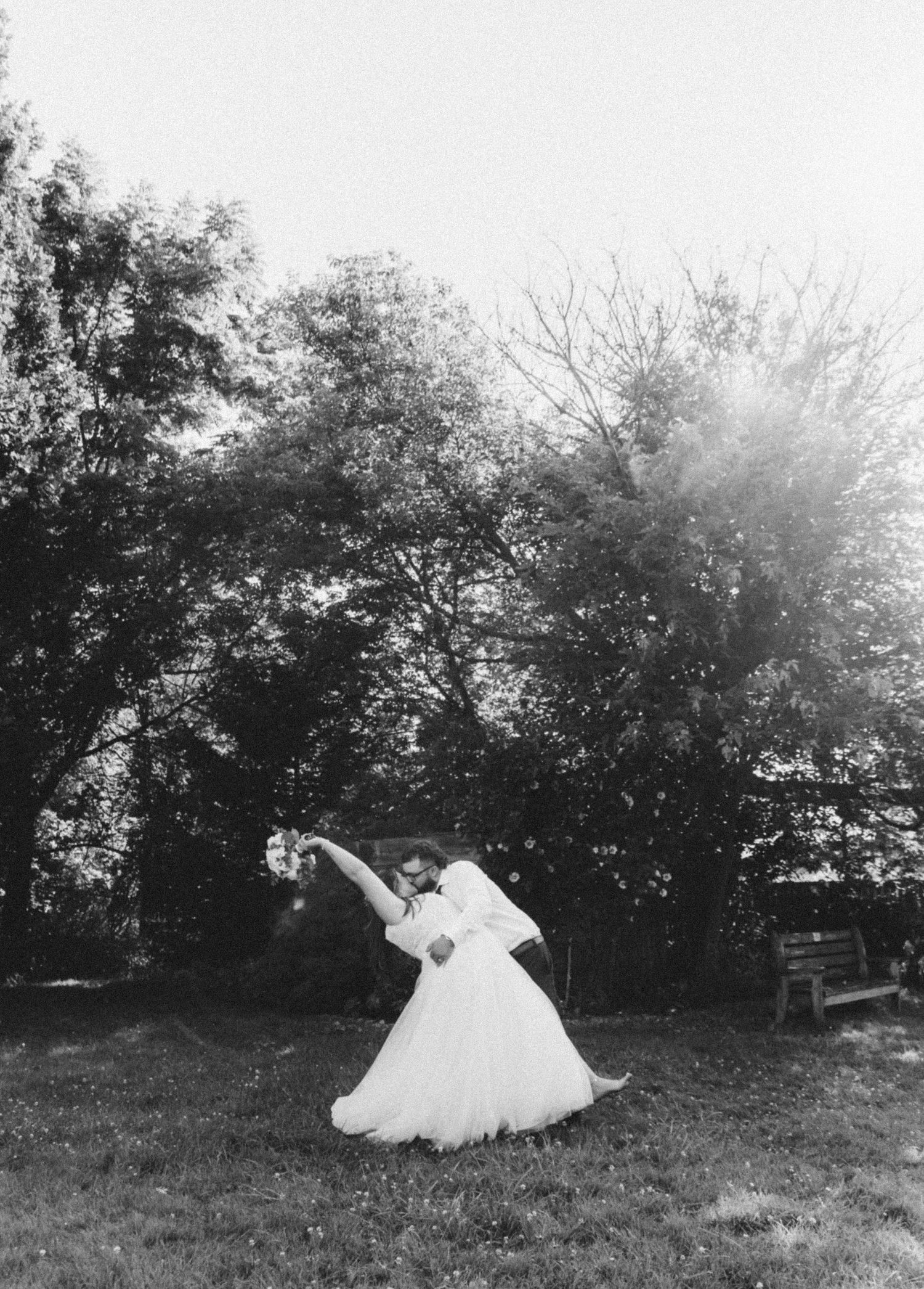 Black and white romantic photo of groom dipping bride in white gown under trees.