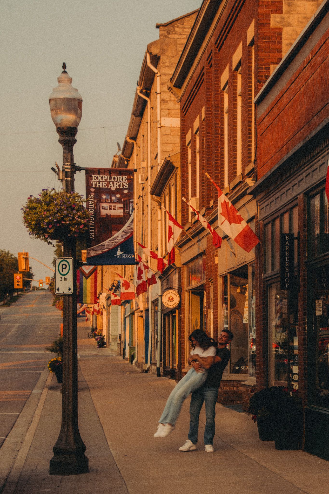 Couple embracing on a charming small-town street lined with Canadian flags and brick buildings at sunset.