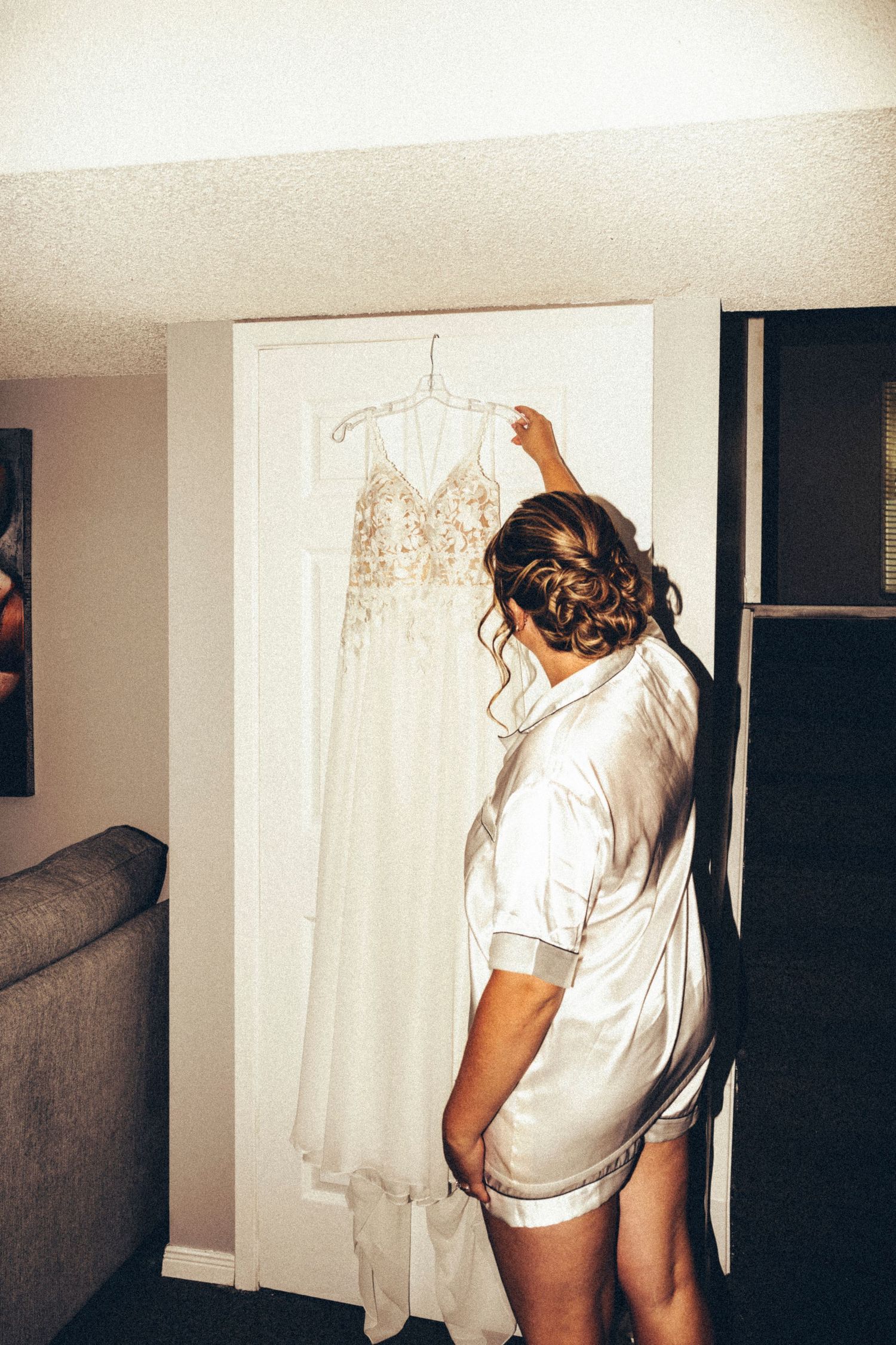 Bride in satin robe admiring her wedding dress hanging in a doorway before getting ready.
