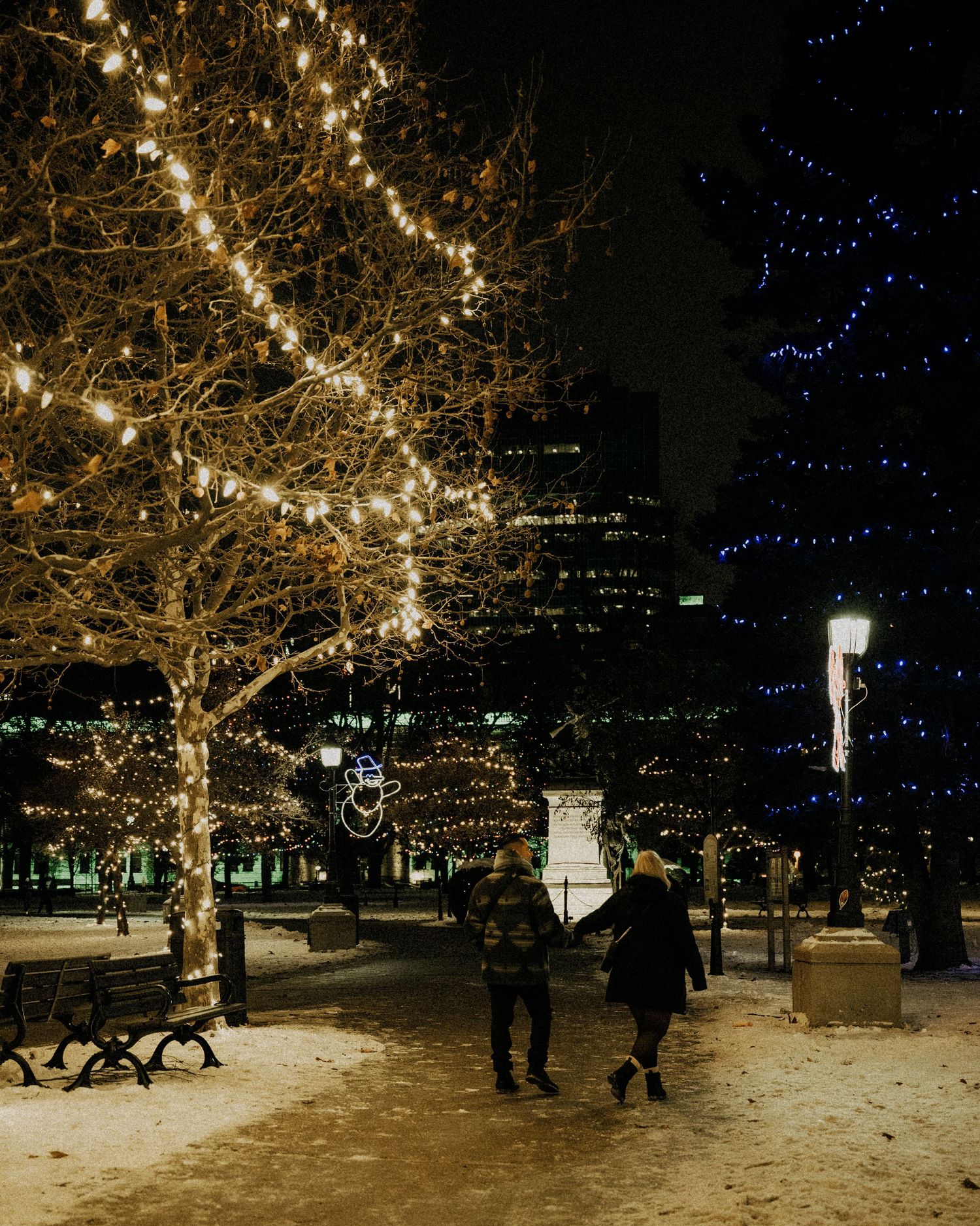 Couple walking through a snowy park decorated with festive holiday lights at night.