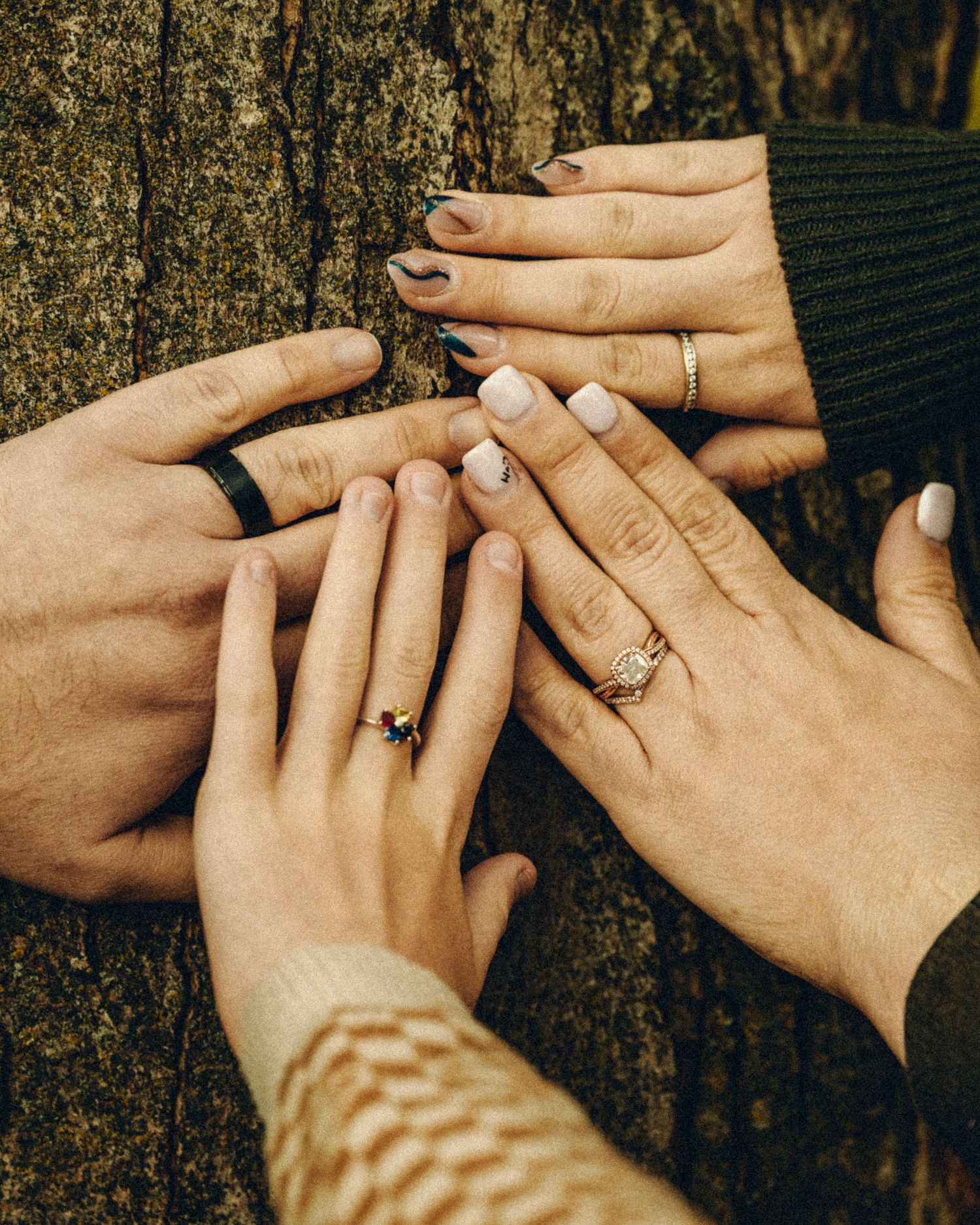 Four hands with rings displayed against a textured tree bark background.