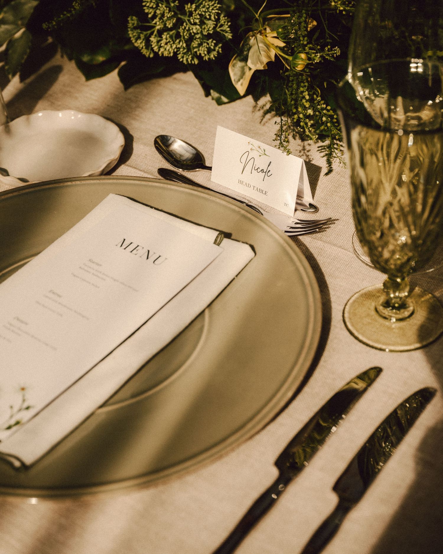 Elegant wedding place setting with menu card, charger plate, crystal glassware and lush greenery centerpiece.