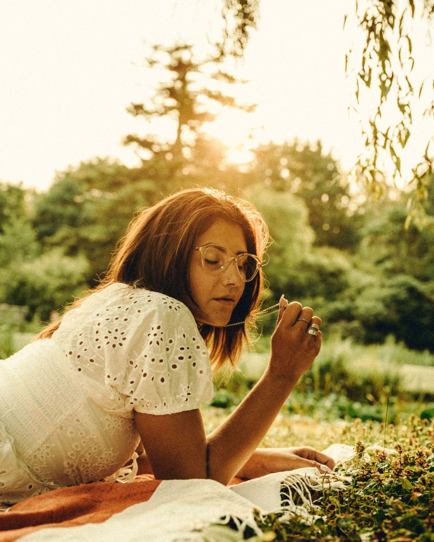 Young woman with glasses in white eyelet dress lying on blanket in golden sunlight.