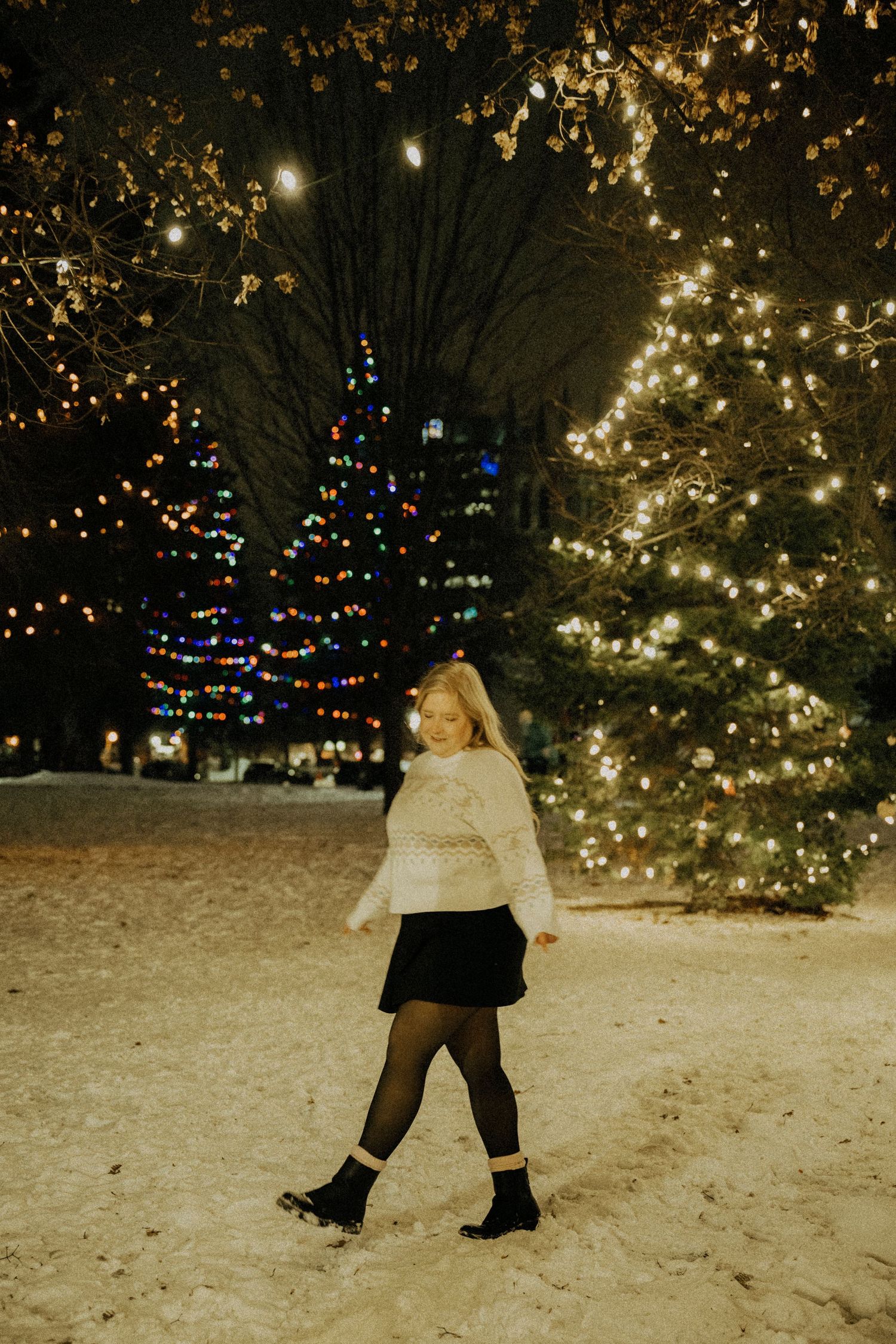 Girl in white sweater and black skirt walking in snowy park with glowing Christmas tree lights at night.