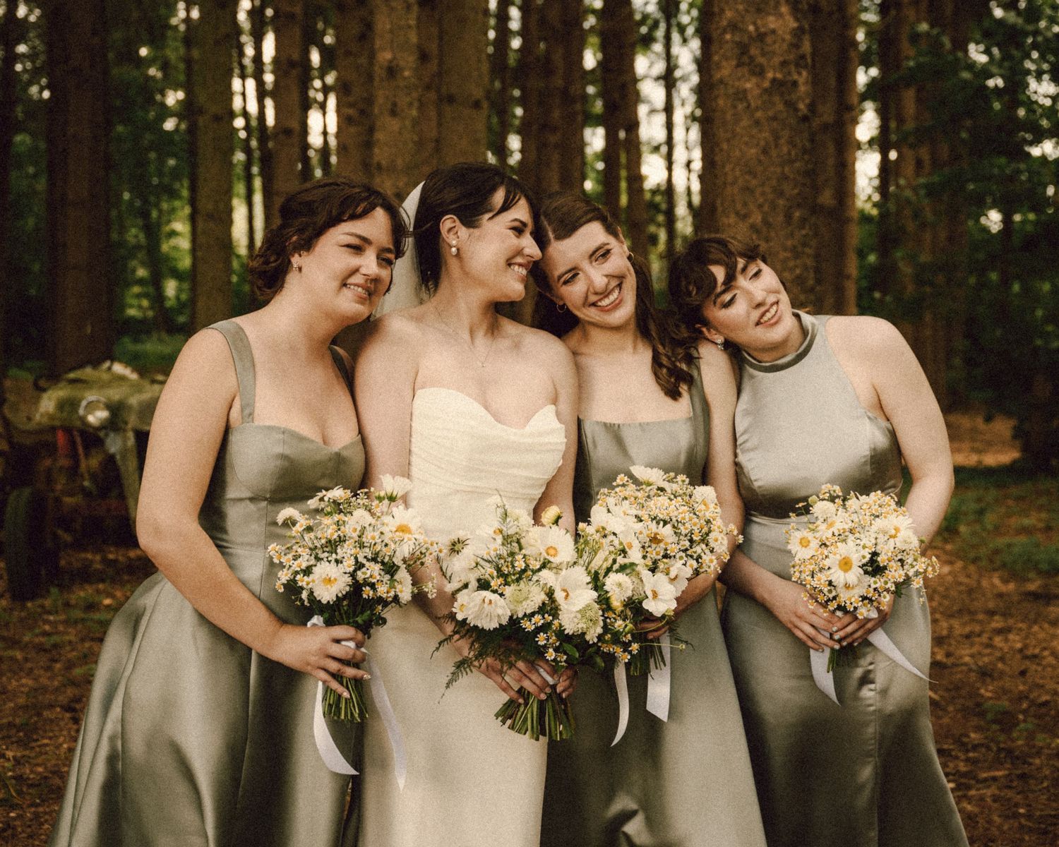 Bride with three bridesmaids in sage green dresses laughing together in a pine forest setting.