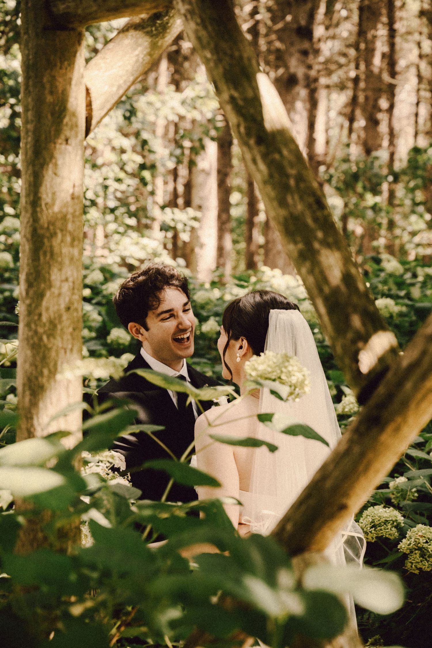 Bride and groom laughing together among lush green foliage and white flowers in a woodland wedding setting.