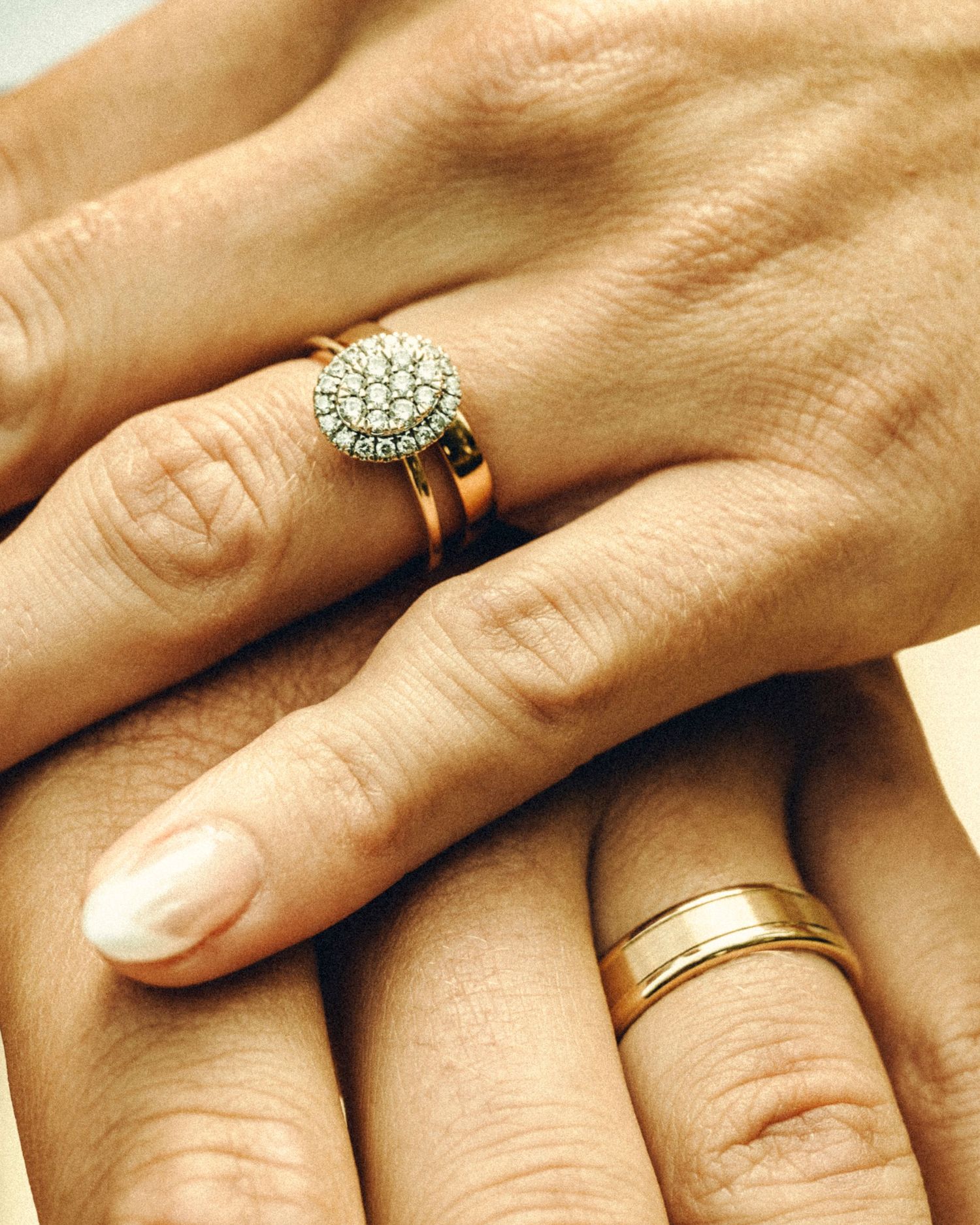 Close-up of bride and groom's hands showing diamond cluster engagement ring and gold wedding bands.