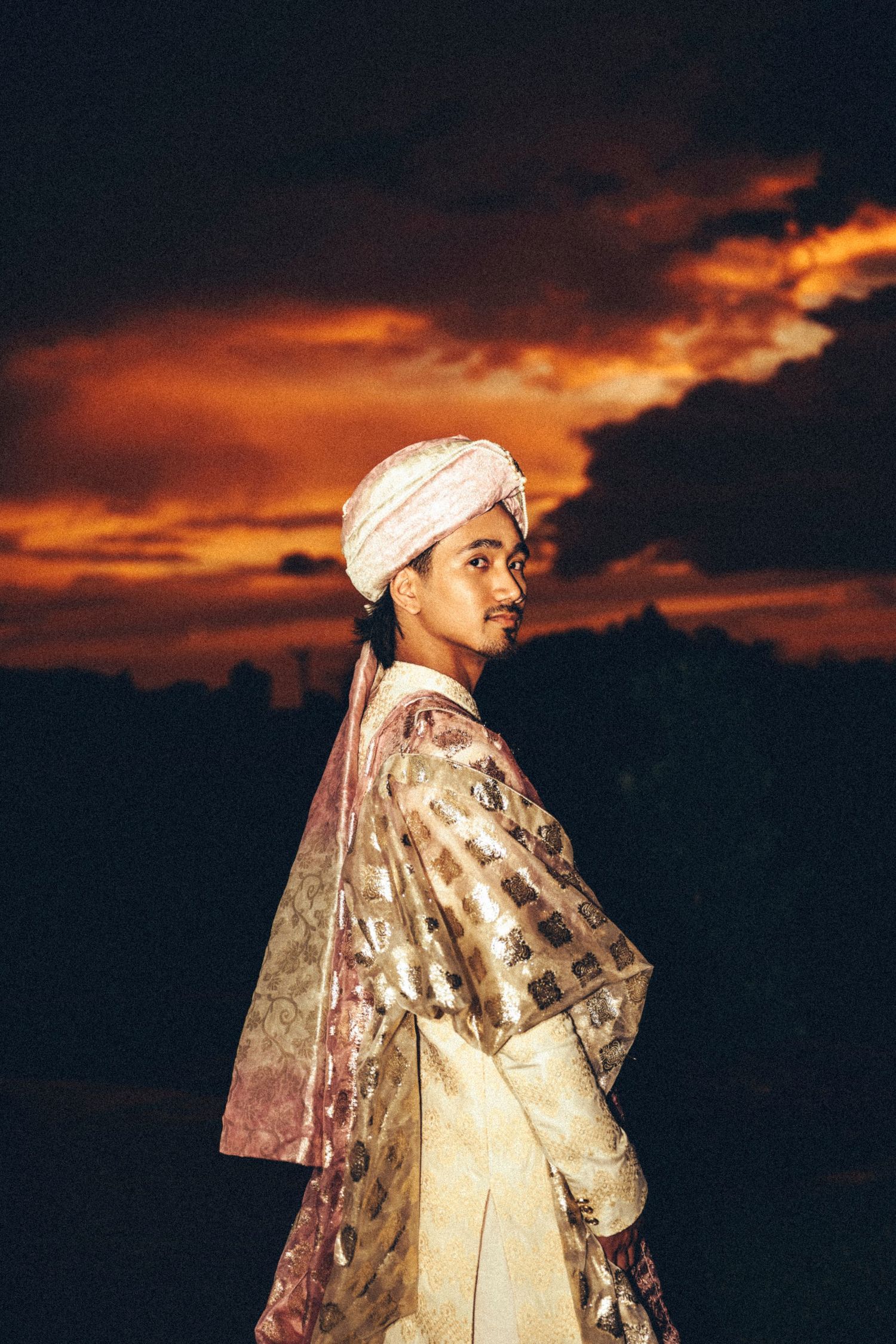 South Asian groom in ivory sherwani and pink turban poses against a dramatic fiery sunset sky.