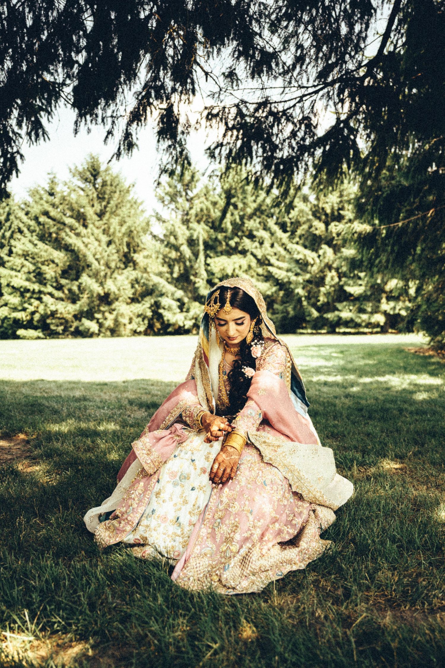 Beautiful South Asian bride in pink and gold lehenga sits gracefully on grass beneath pine trees in an outdoor setting.