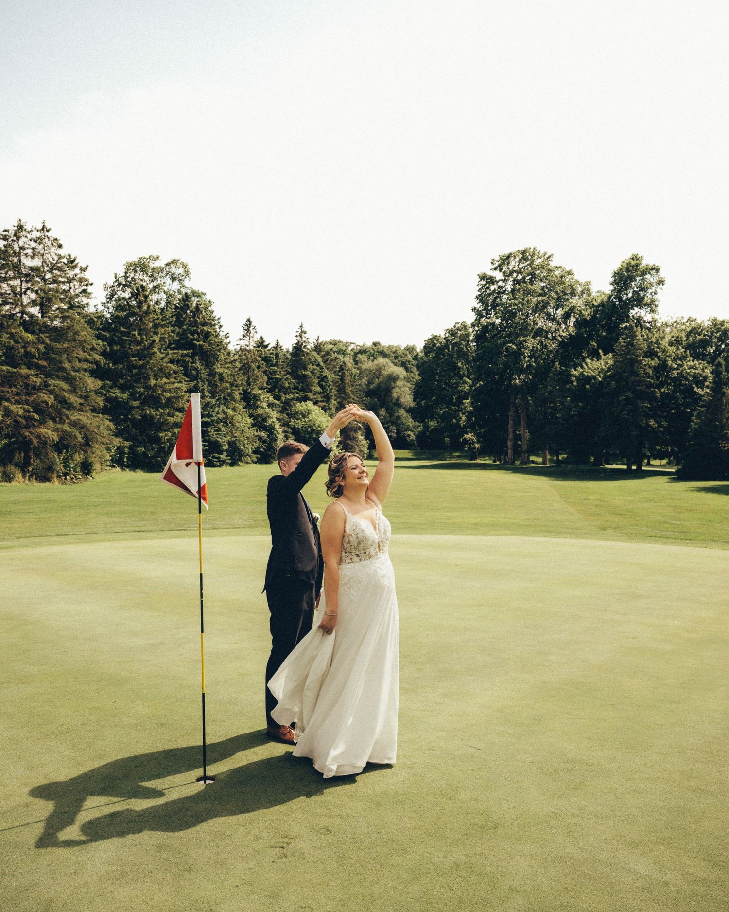 Bride and groom dance together on a golf course green beside a red flag on sunny day.
